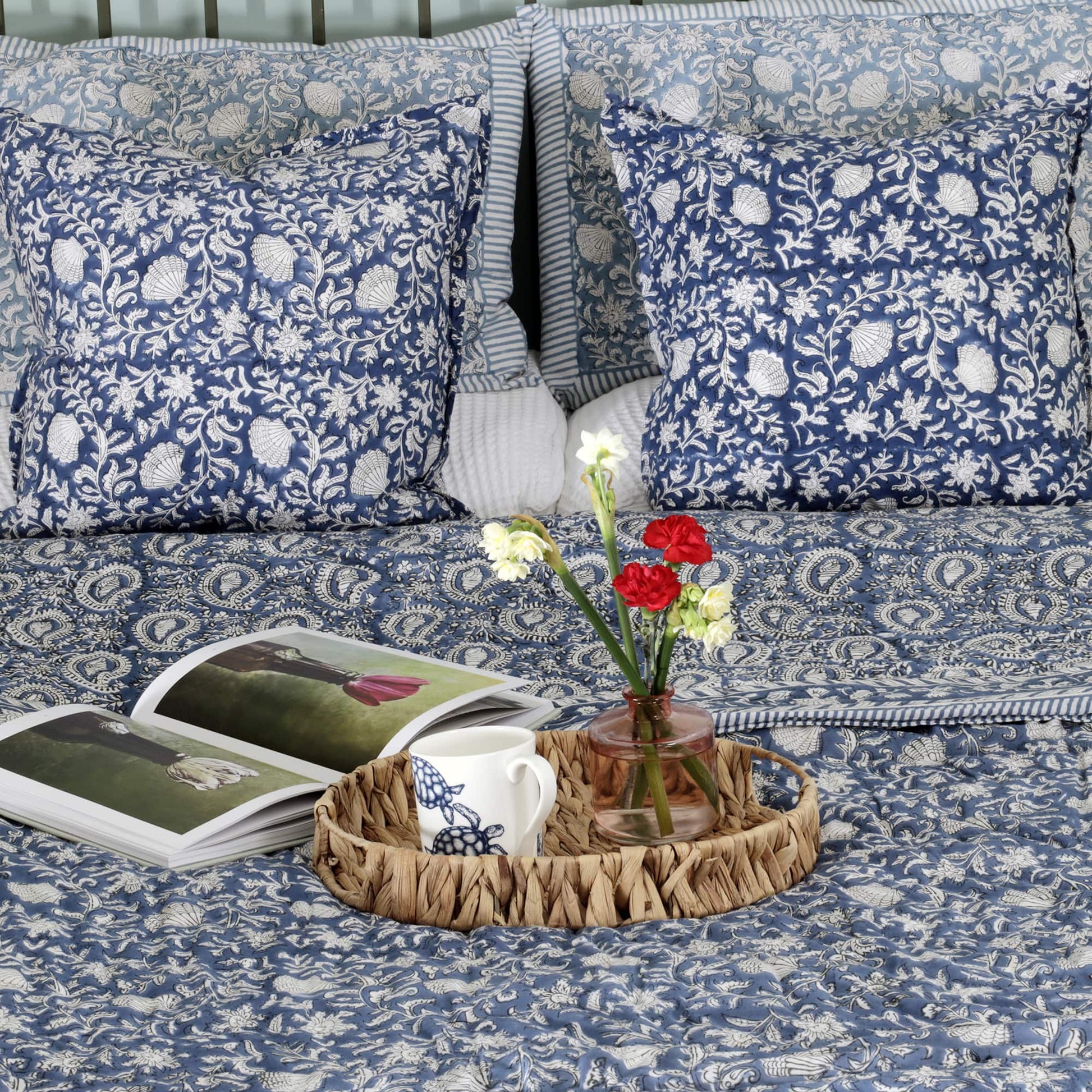 A cozy bedroom setup featuring blue and white block-printed cushions with floral and shell patterns, layered with a paisley-patterned quilt. A woven tray placed on the bed holds a vase with red and white flowers, a mug, and an open book with nature photography. The setting exudes a warm and inviting ambiance.