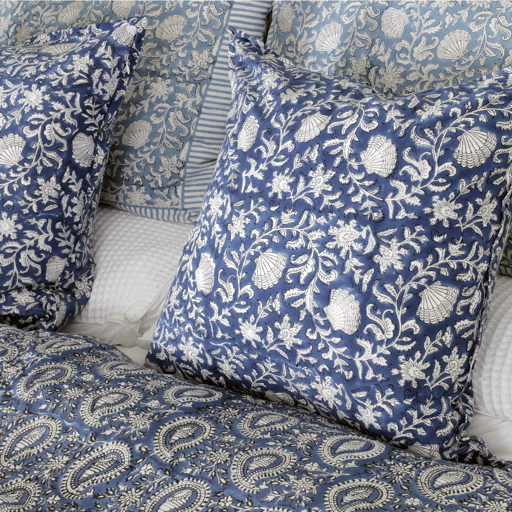 A bedroom setting featuring blue and white cushion covers with intricate floral and shell patterns. The cushions are paired with a quilted paisley-patterned bedspread and additional striped and floral pillows in the background. The overall design conveys a coordinated and elegant look.