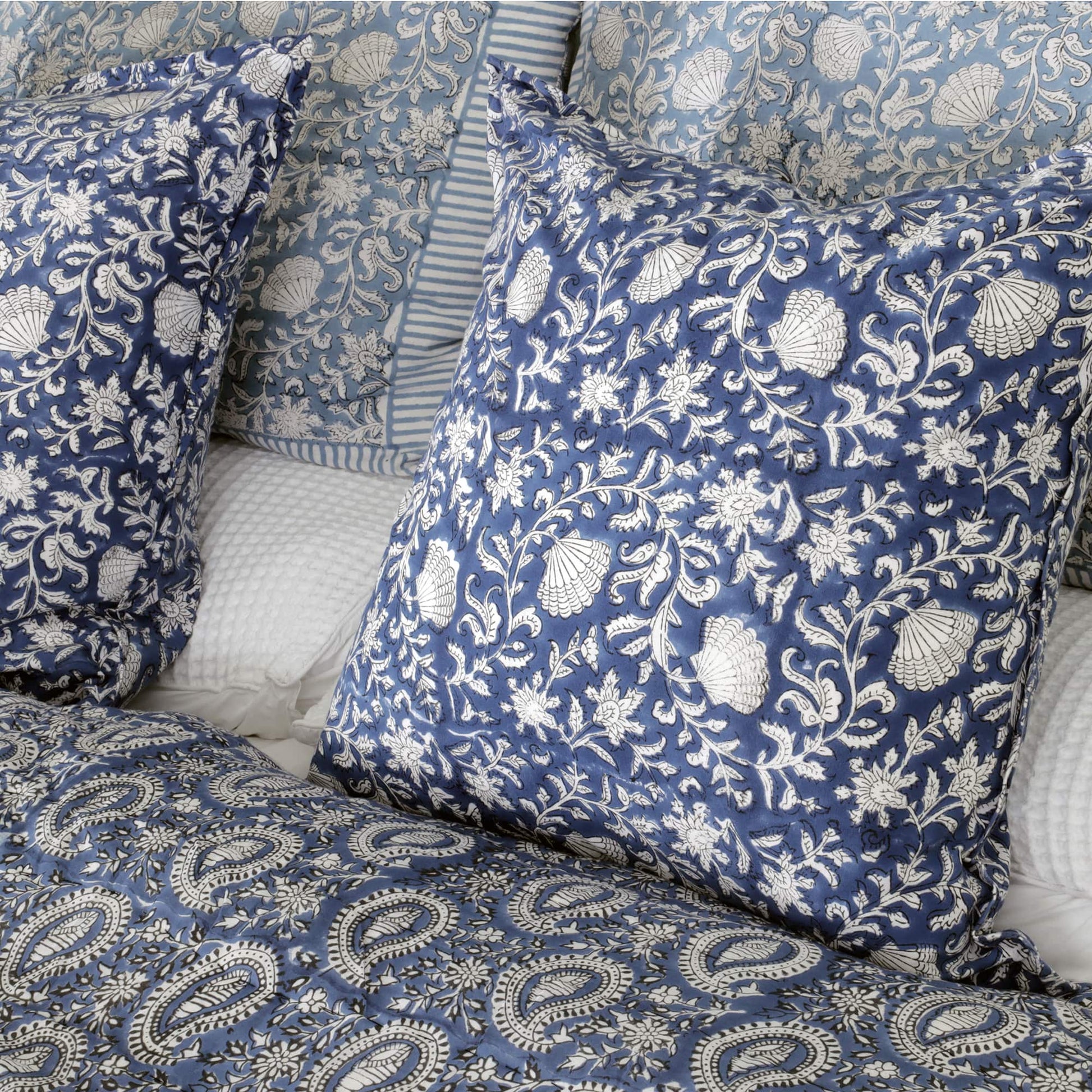 A bedroom setting featuring blue and white cushion covers with intricate floral and shell patterns. The cushions are paired with a quilted paisley-patterned bedspread and additional striped and floral pillows in the background. The overall design conveys a coordinated and elegant look.