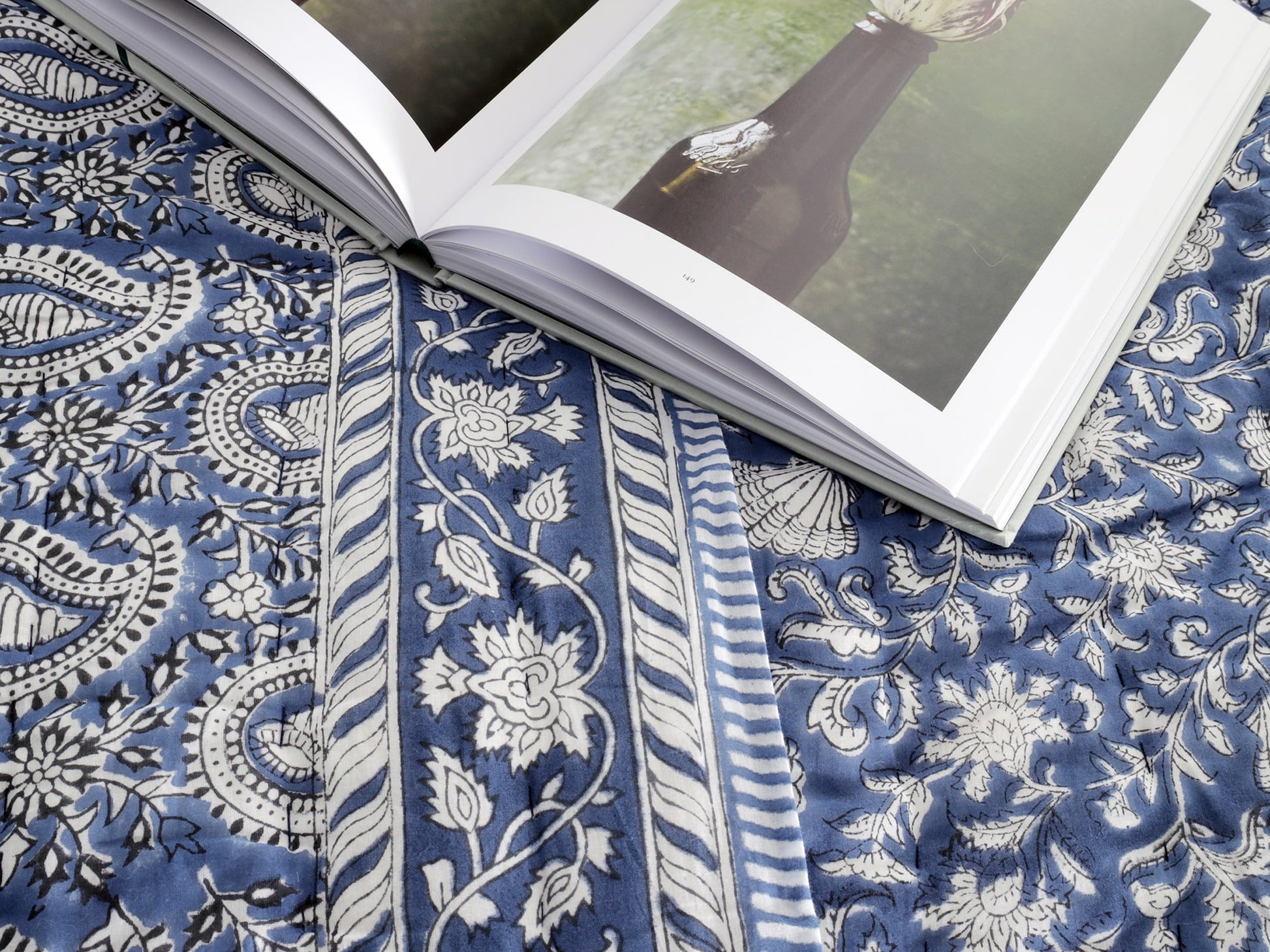 Close-up of an open book placed on a blue quilt with intricate paisley, floral, and seashell patterns. The detailed block print design of the quilt complements the artistic imagery in the book.
