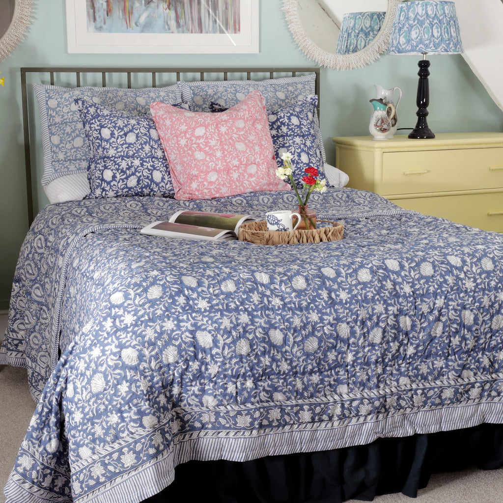 A stylish bedroom arrangement featuring a bed dressed in blue and white block print bedding with intricate floral patterns. A single pink cushion with matching block print design adds a pop of color at the center of the bed. The setup includes a wicker tray with a cup and flowers, a yellow dresser, and decorative elements like a framed artwork and patterned lampshades, creating a charming and cozy atmosphere.