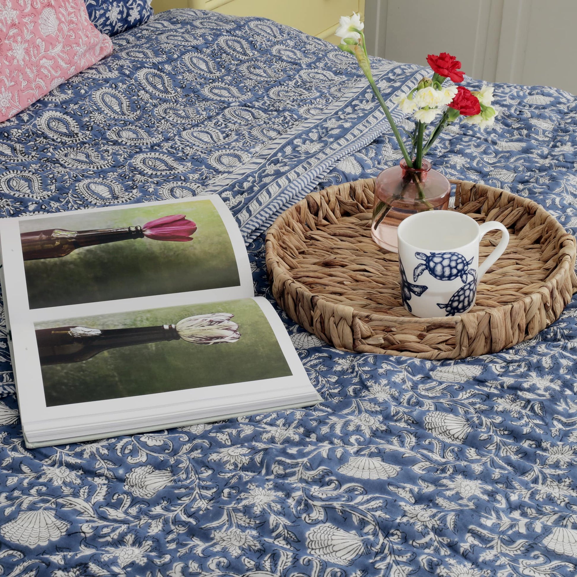 Cozy bedroom scene featuring a blue quilt with intricate paisley and floral patterns. An open book with botanical artwork rests on the bed alongside a wicker tray holding a mug with a turtle design, a small vase with red and white flowers, and a warm, inviting aesthetic.