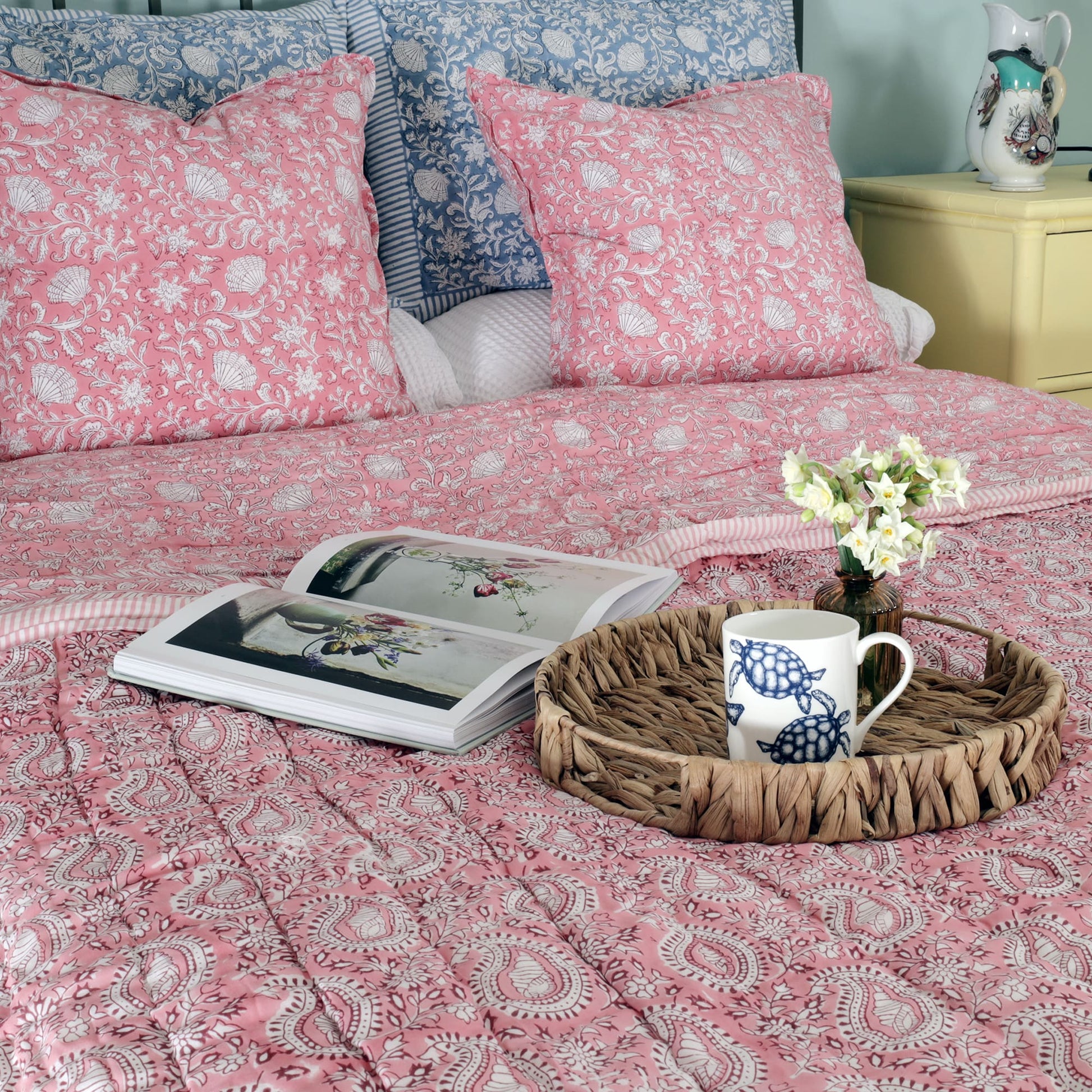 A cozy bedroom scene featuring a bed adorned with pink block print bedding with intricate floral and paisley patterns. Two matching pink cushions rest against blue floral block print pillows in the background. On the bed, a wicker tray holds a mug with a turtle design and a small vase with white flowers, alongside an open book displaying floral artwork. A yellow dresser with decorative items completes the inviting setup.