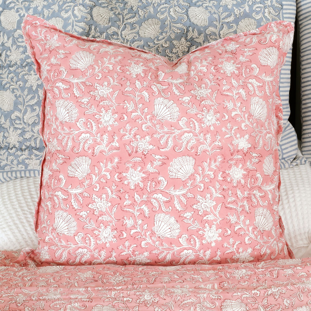 A pink square cushion with white floral and seashell block print patterns, placed on a bed layered with coordinating bedding in soft blues and whites. The cushion adds a vibrant, elegant touch to the cozy setup.