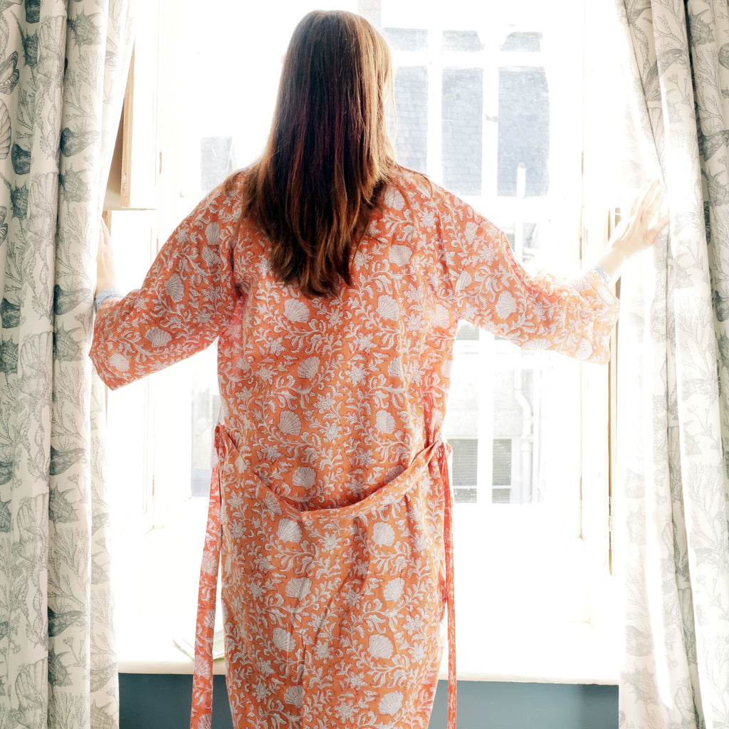 A woman wearing a coral-colored dressing gown with a white floral and seashell block print design, standing by a bright window with her hands parting the curtains, her back to the camera.