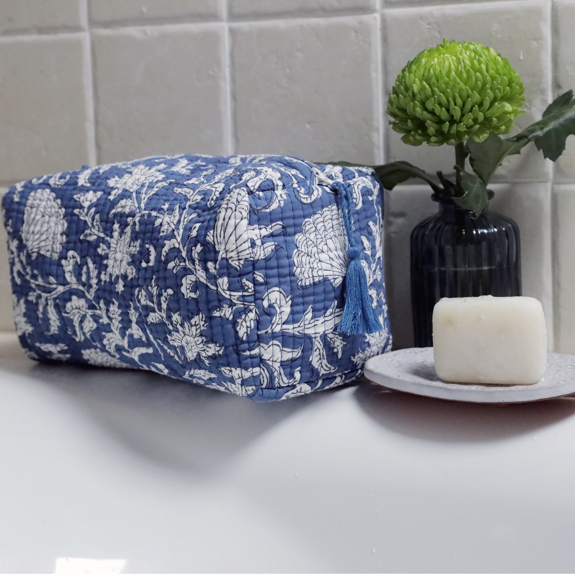 A blue and white quilted toiletry bag with floral patterns sits on a white bathroom counter. A green chrysanthemum flower in a dark ribbed glass vase is placed behind the bag, and a white bar of soap rests on a round dish in the foreground.