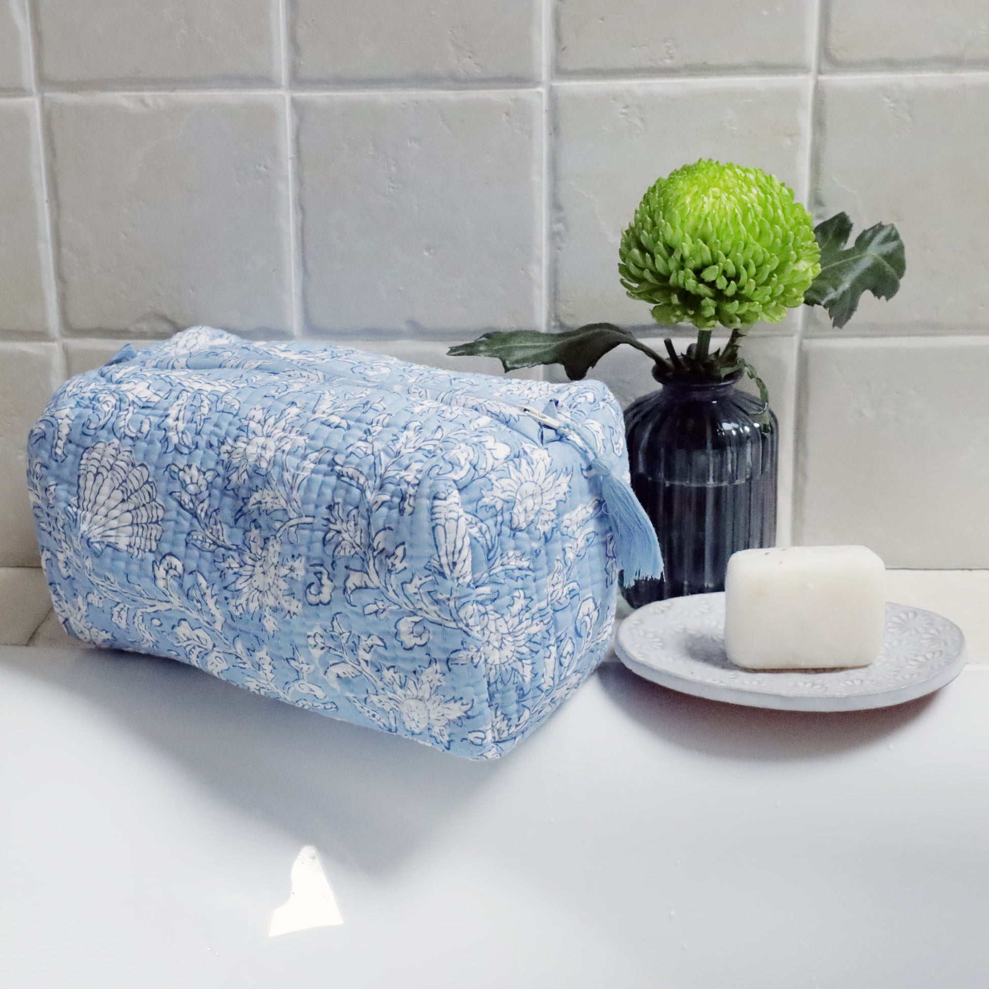 Light blue quilted washbag with intricate white floral and seashell block print patterns, placed on the edge of a white sink. The scene is complemented by a small vase with a vibrant green flower and a soap bar resting on a decorative dish, creating a fresh and elegant bathroom setup.