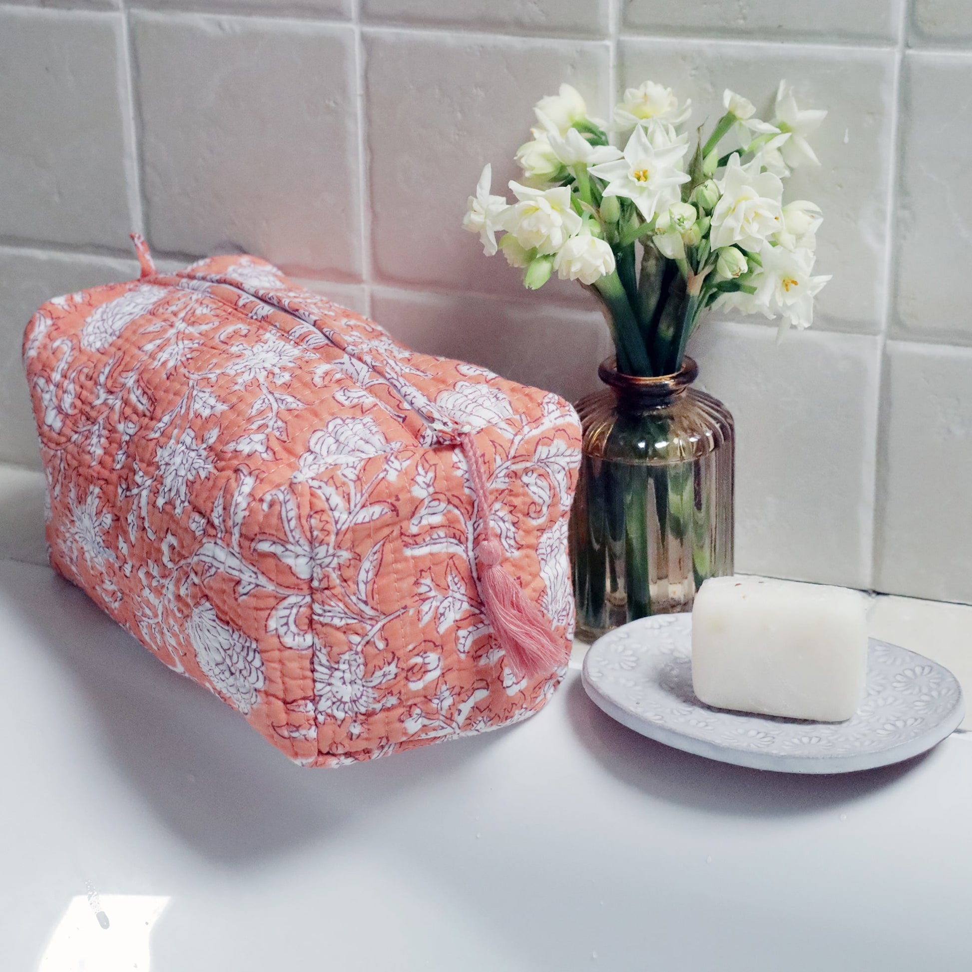 A coral-colored quilted wash bag with a white floral and seashell block print design, placed on a bathroom counter beside a small vase of white flowers and a soap bar on a decorative dish, set against a tiled wall.