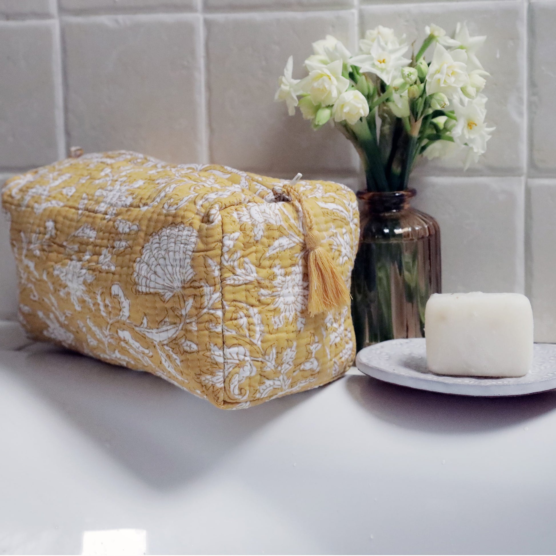 Yellow quilted washbag featuring intricate white floral and seashell block print patterns, placed on the edge of a white sink. The scene is complemented by a small vase with white flowers and a soap bar resting on a decorative dish, creating a fresh and elegant bathroom setup.
