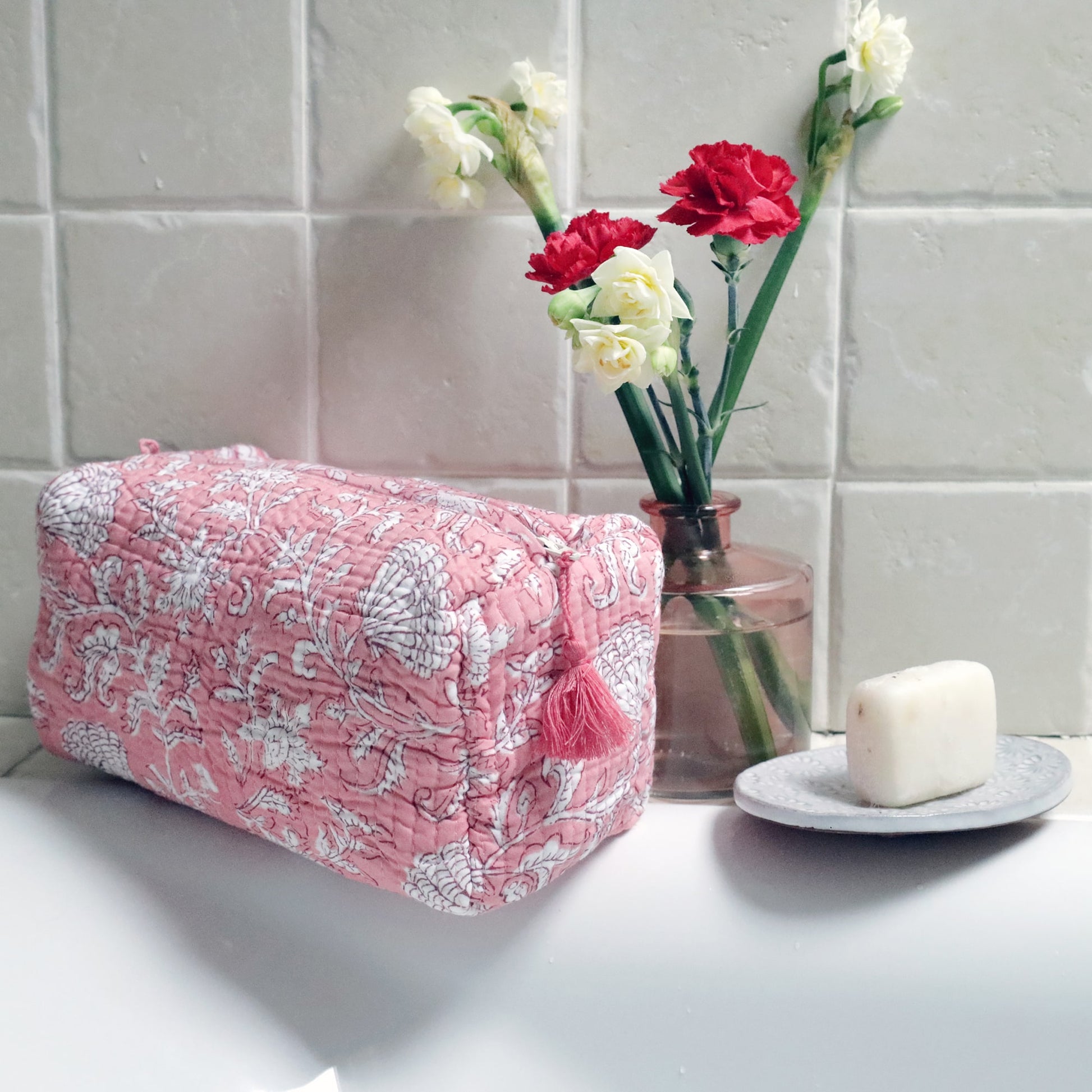 A pink quilted wash bag featuring white floral and seashell block print patterns with a pink tassel on the zipper, placed on the edge of a white bathtub. Next to it, a vase with red and cream flowers and a soap dish with a white bar of soap sit against a tiled wall.
