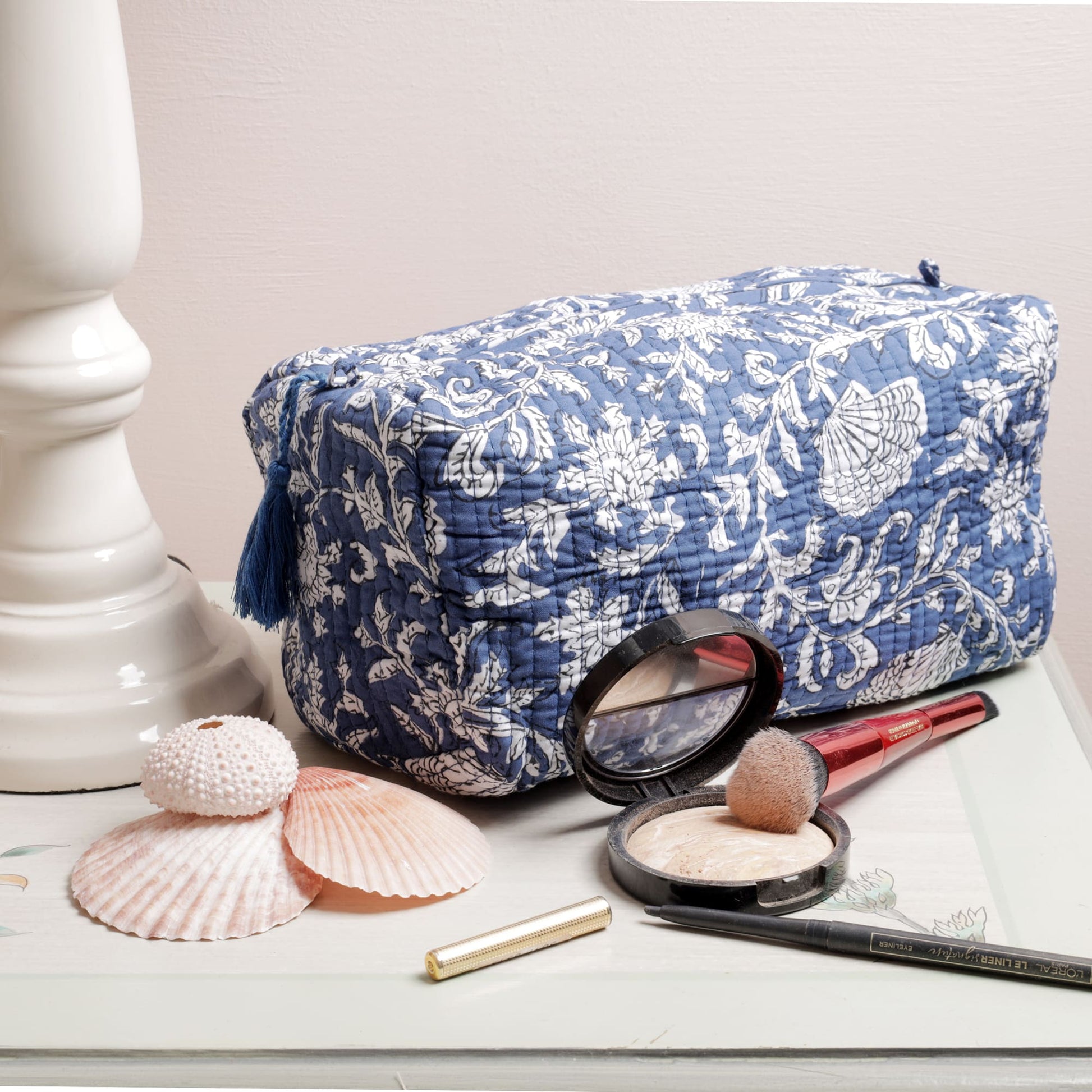Quilted blue and white floral pouch with a tassel zipper displayed on a decorative surface. Surrounding it are seashells and beauty items, including a makeup brush, compact powder, eyeliner, and a lipstick, creating a stylish and functional vanity setup.