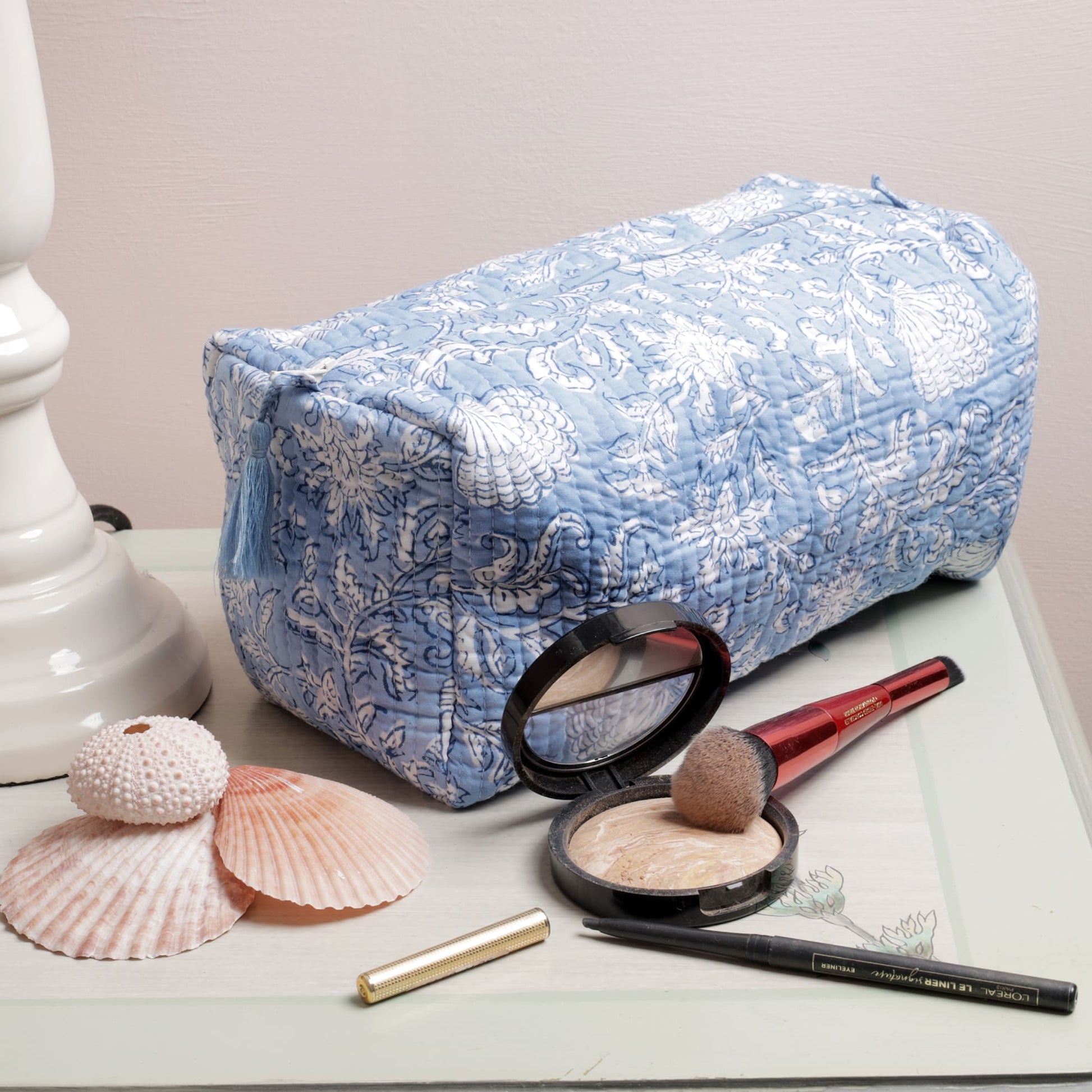 Light blue quilted washbag with intricate white floral block print patterns, displayed on a painted surface. The setup includes seashell decorations and makeup items such as a compact powder, brush, eyeliner, and lipstick, adding a functional and stylish touch.