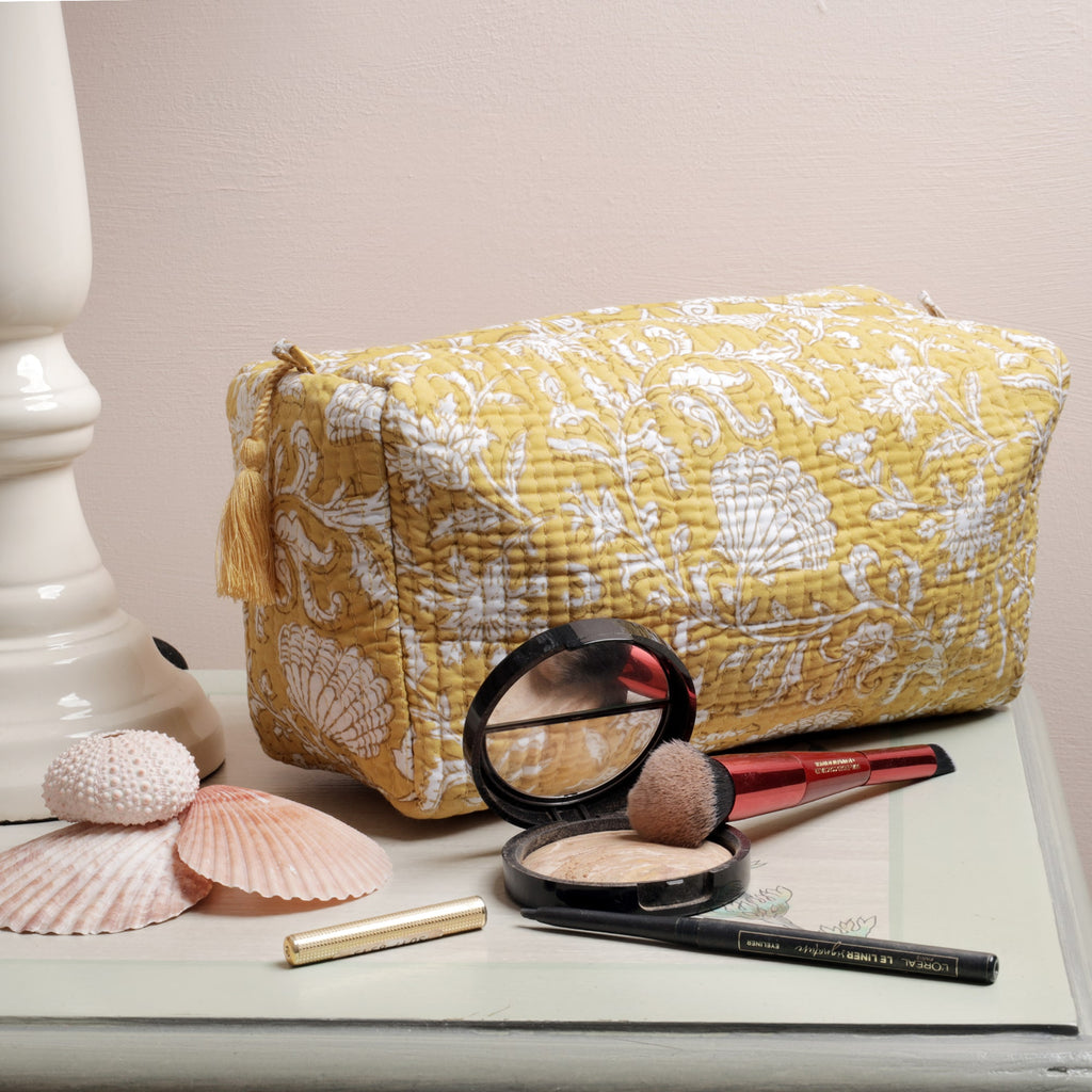 Yellow quilted washbag featuring intricate white floral and seashell block print patterns, displayed on a painted surface. The setup includes seashell decorations and makeup items such as a compact powder, brush, eyeliner, and lipstick, adding a functional and stylish touch.