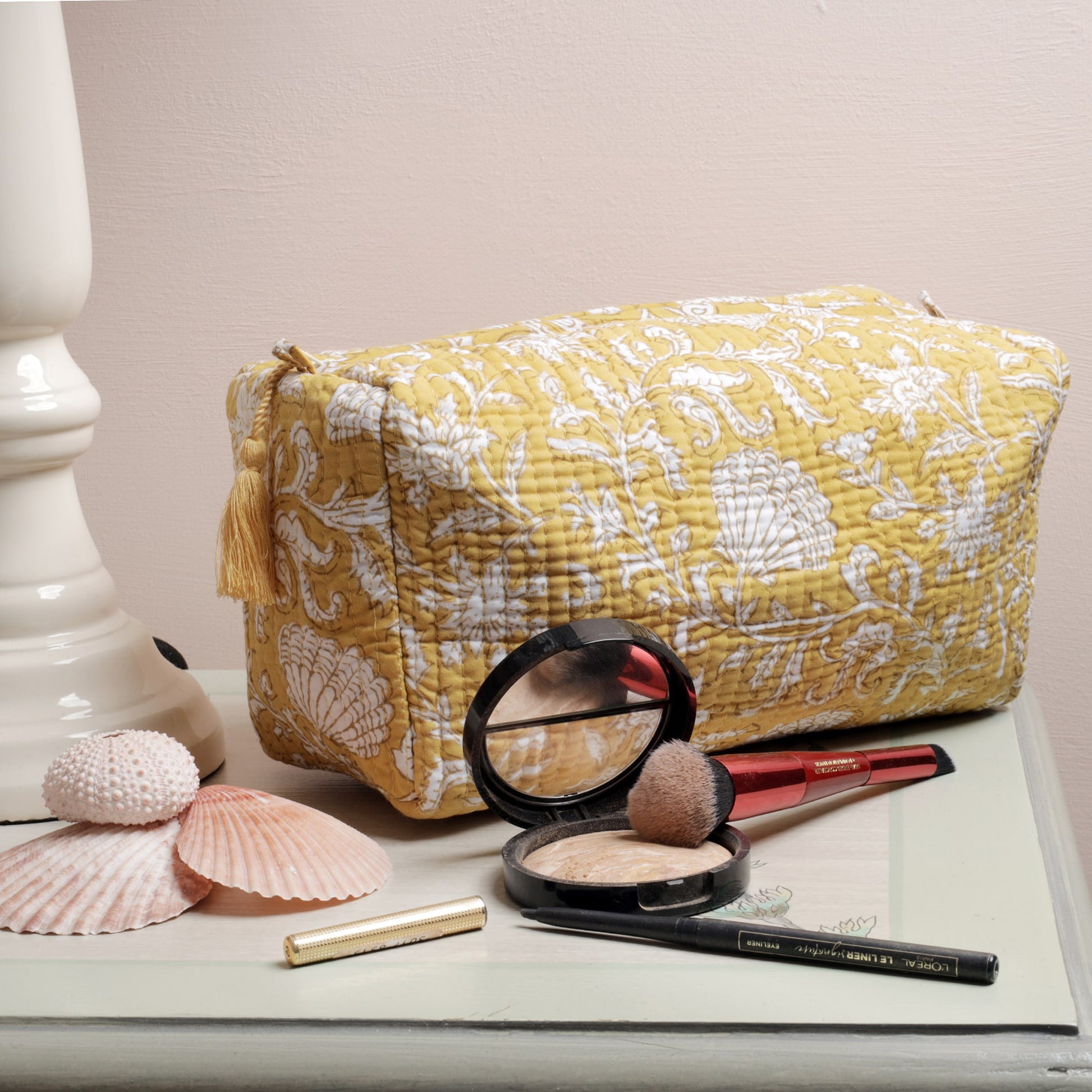 Yellow quilted washbag featuring intricate white floral and seashell block print patterns, displayed on a painted surface. The setup includes seashell decorations and makeup items such as a compact powder, brush, eyeliner, and lipstick, adding a functional and stylish touch.