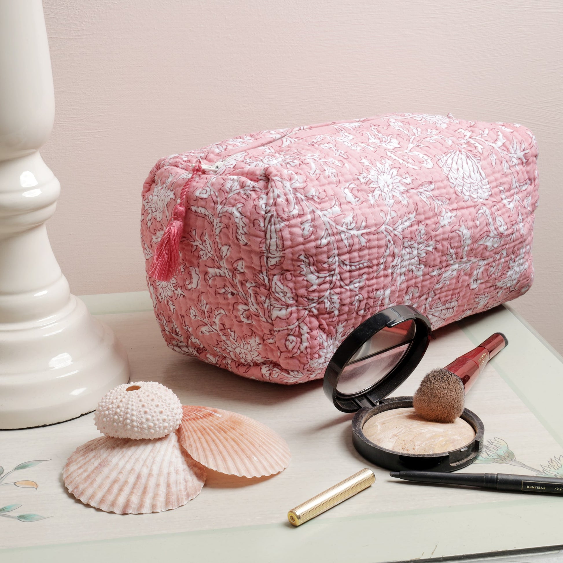 A pink quilted wash bag with white floral and seashell block print patterns, adorned with a pink tassel on the zipper, displayed on a light-colored table alongside seashells, makeup items including a brush, compact powder, and eyeliner