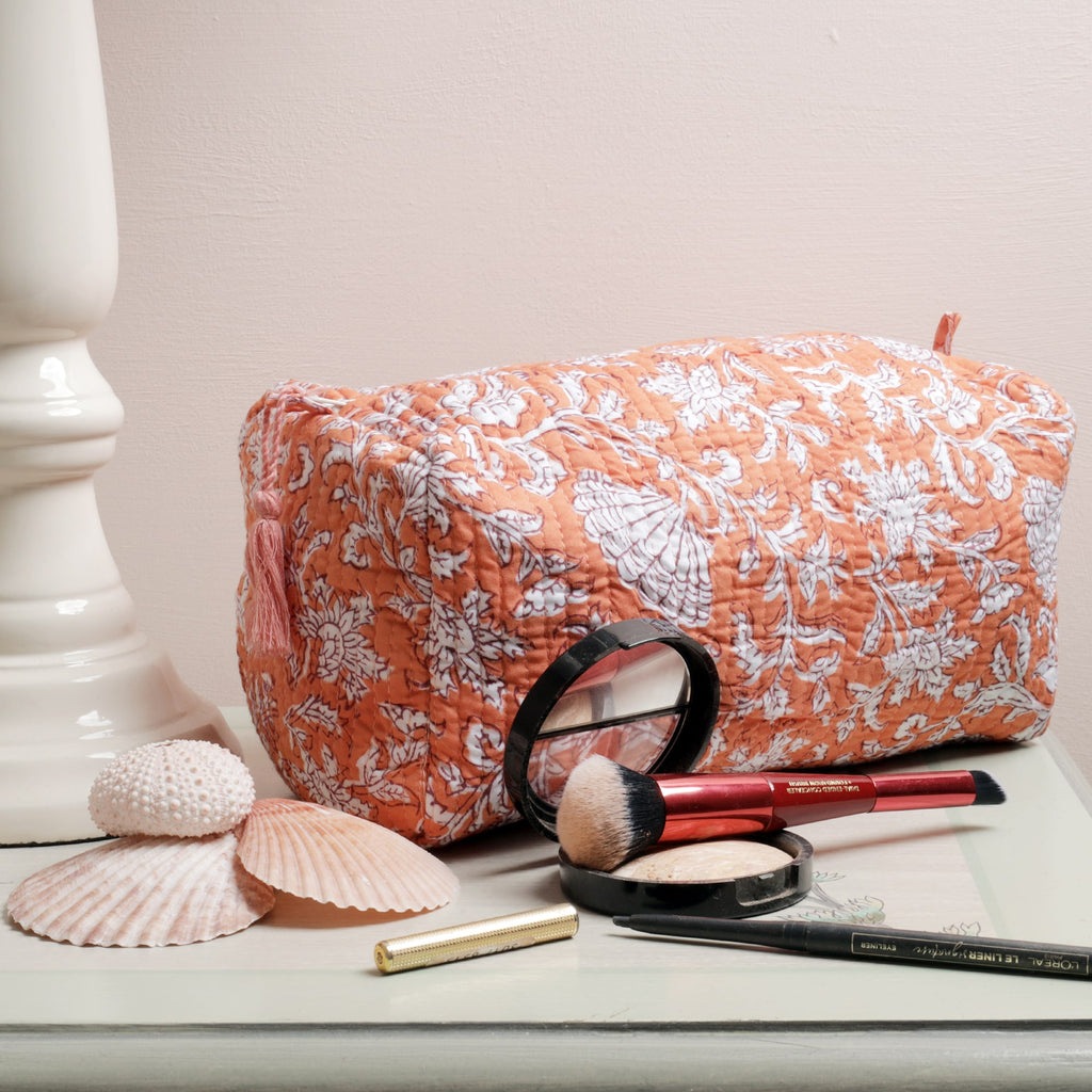 A coral-colored quilted wash bag with a white floral and seashell block print design, placed on a light-colored surface next to seashell decorations, makeup brushes, compact powder, eyeliner, and lipstick.