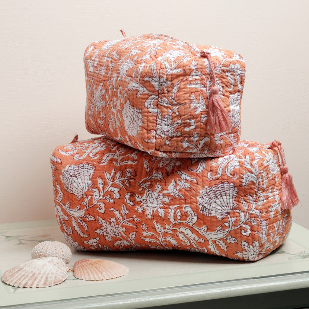 Two coral-colored quilted wash bags with white floral and seashell block print designs, stacked on a light-colored surface, accompanied by decorative seashells in the foreground.