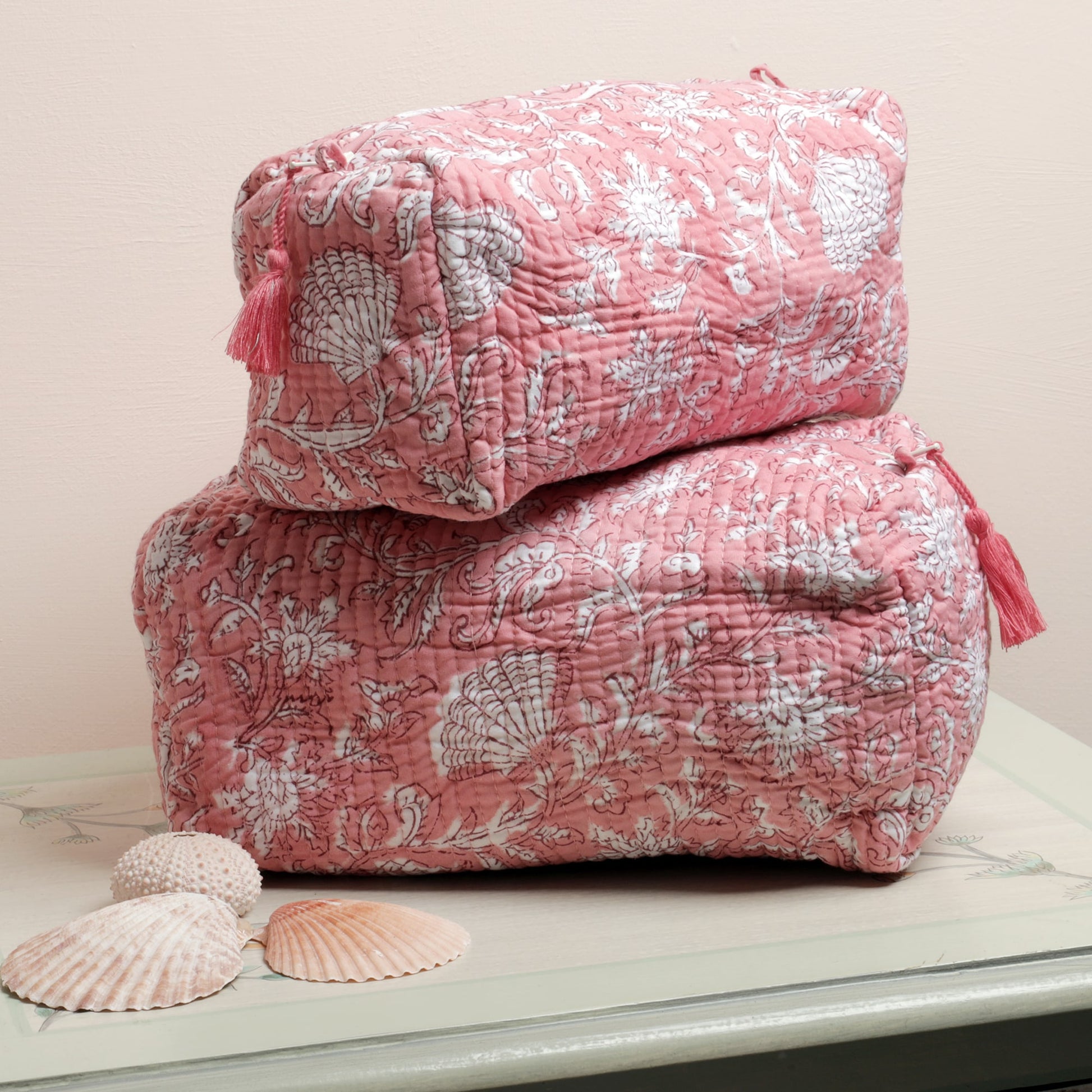 Two pink quilted wash bags with intricate block print designs featuring white floral and seashell patterns, adorned with pink tassels on the zippers, stacked on a light-colored table next to seashell decor.