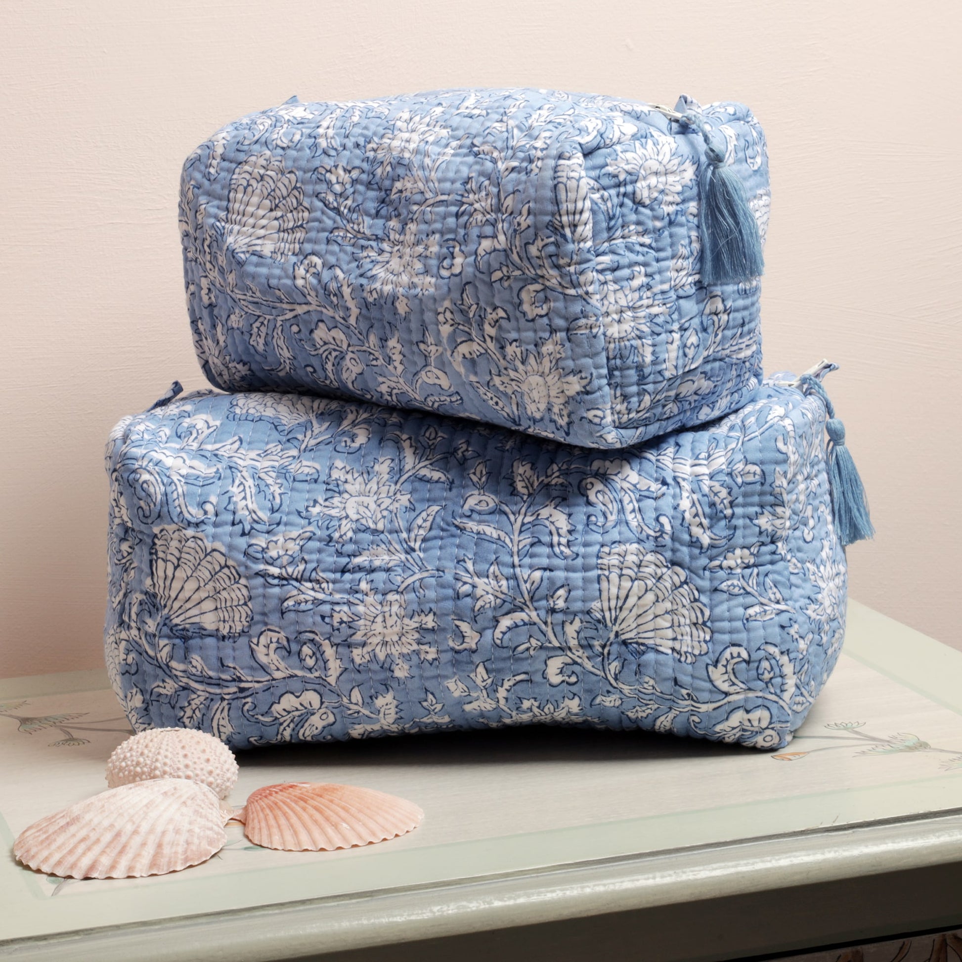 Two stacked light blue quilted washbags featuring intricate white floral and seashell block print patterns. Each bag includes a tassel detail on the zipper. The setup is complemented by seashell decorations placed on a painted surface, creating a coastal-inspired look
