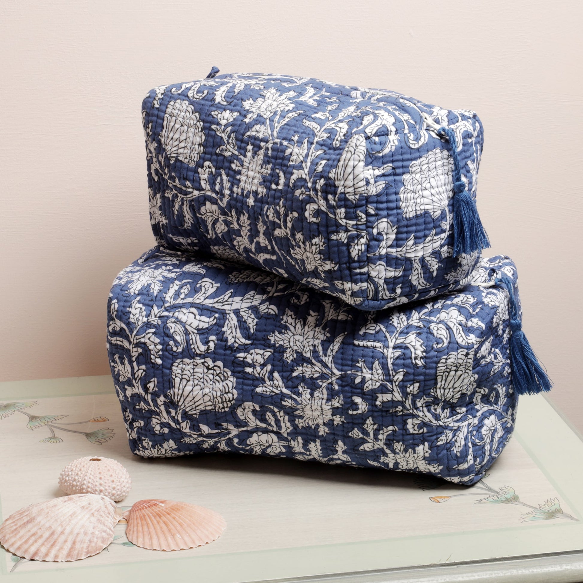 Two quilted rectangular pouches with a blue and white floral pattern stacked on a decorative surface. Each pouch features a zippered closure with a matching blue tassel. The setup includes seashell accents, adding a coastal, elegant touch to the display.