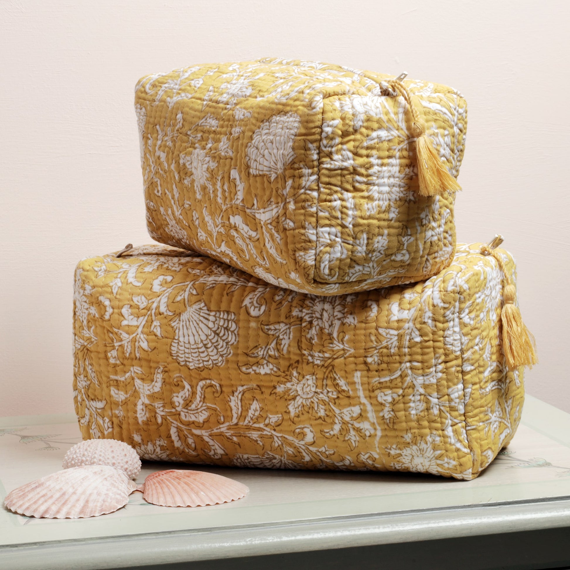 Two stacked yellow quilted washbags featuring intricate white floral and seashell block print patterns. Each bag includes a tassel detail on the zipper, complemented by seashell decorations placed on a painted surface, creating a coastal-inspired aesthetic.