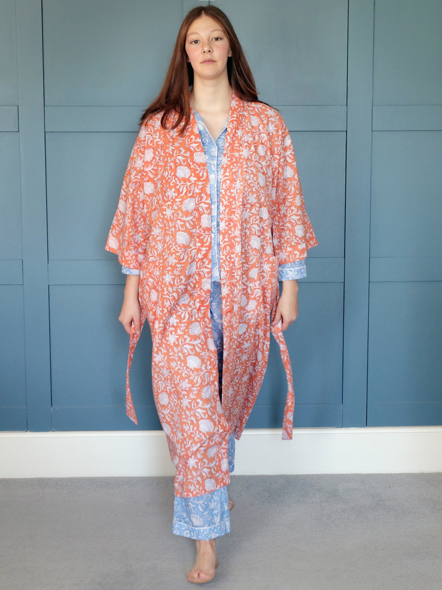A young woman standing barefoot on a light carpet in front of a blue-paneled wall, wearing a coral block print dressing gown with white floral and seashell patterns over light blue pajamas with intricate designs.