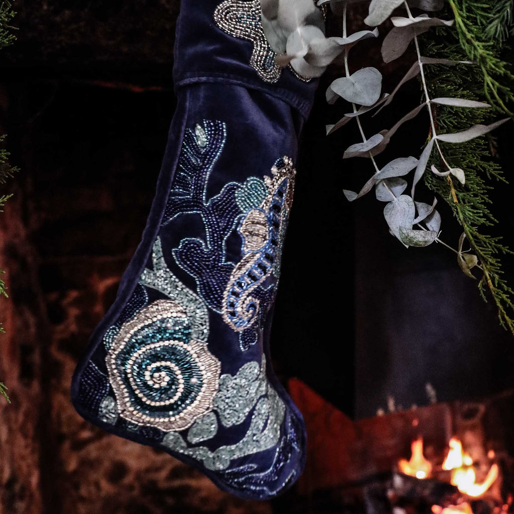 dark blue velvet Christmas stocking decorated with blue beads and silver beads with corals, seaweed starfish, ammonite and seahorse hanging on foliage covers mantlepiece above a roaring fire.