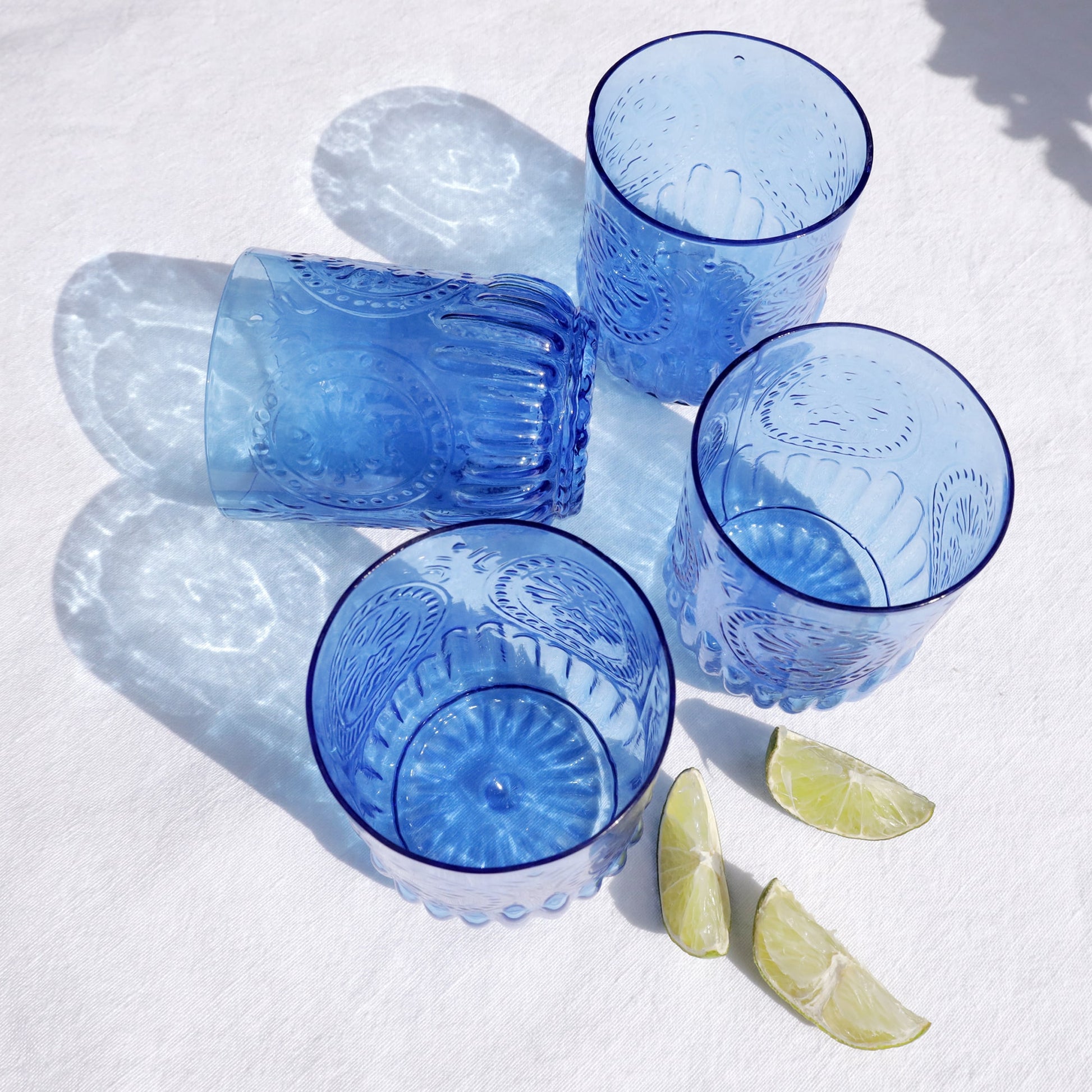Set of four embossed blue glass tumblers arranged on a white surface with sunlight casting reflections, accompanied by lime wedges.