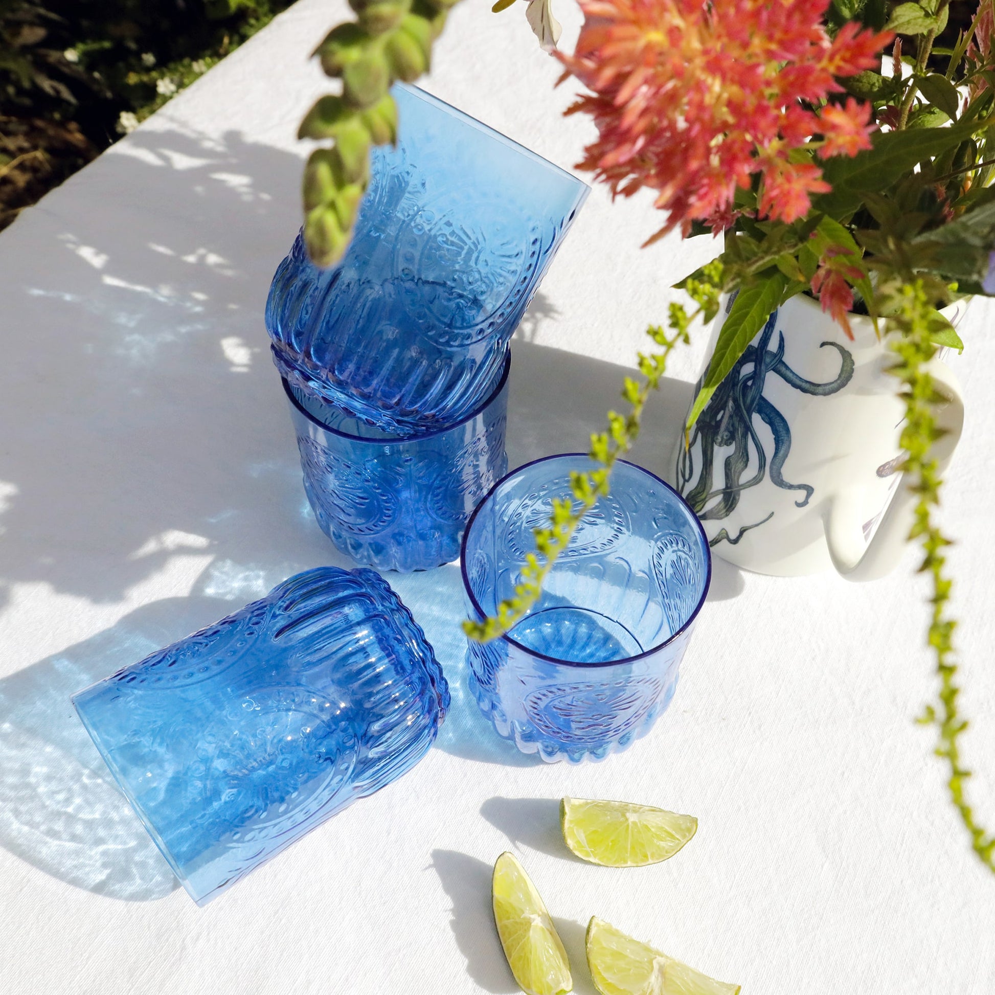 Set of embossed blue glass tumblers arranged on a white surface with lime wedges, beside a white jug decorated with a blue octopus illustration and filled with vibrant flowers.