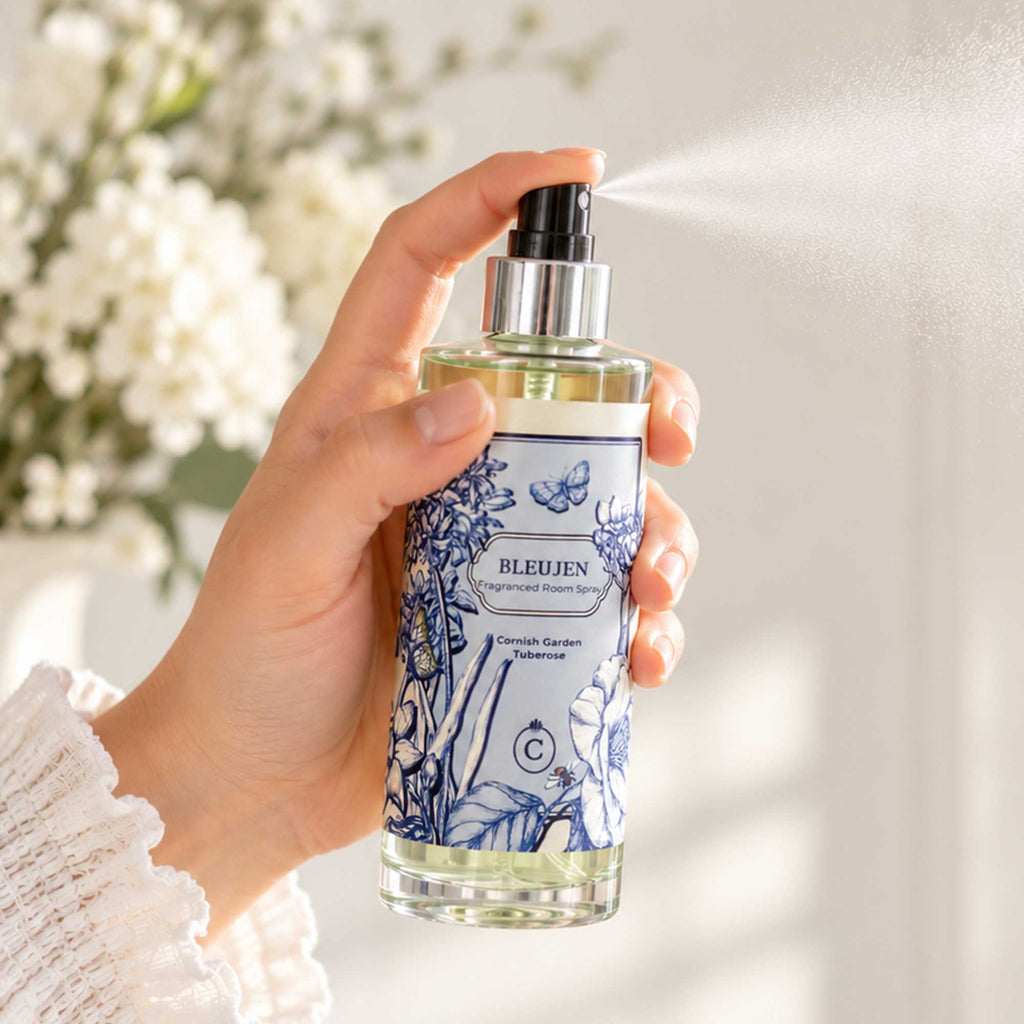 Alt text: Cream Cornwall Bleujen fragranced room spray being sprayed in a bright, softly styled interior, with a fine mist and white flowers in the background.