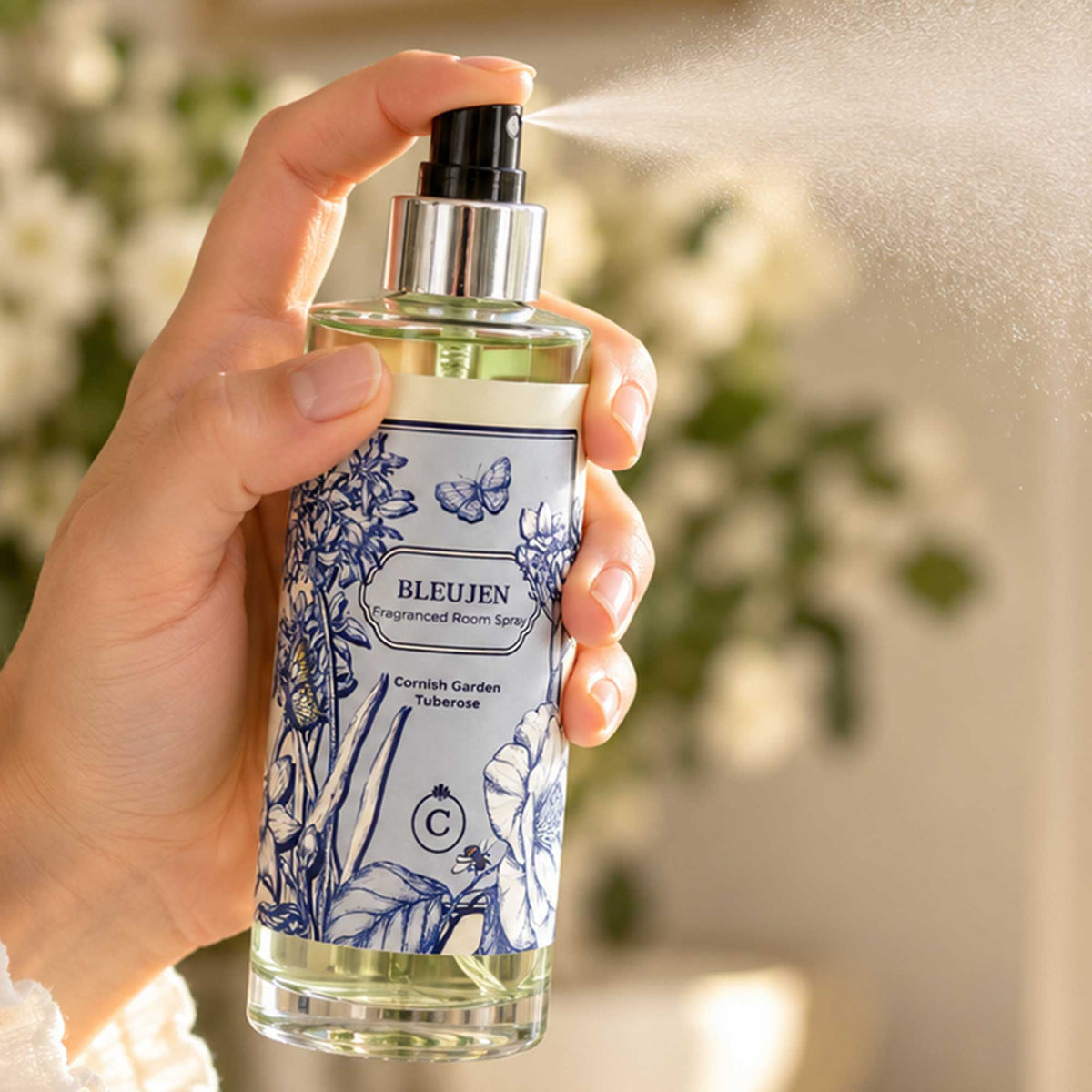 Cream Cornwall Bleujen fragranced room spray being sprayed in a bright, softly styled interior, with a fine mist and white flowers in the background.