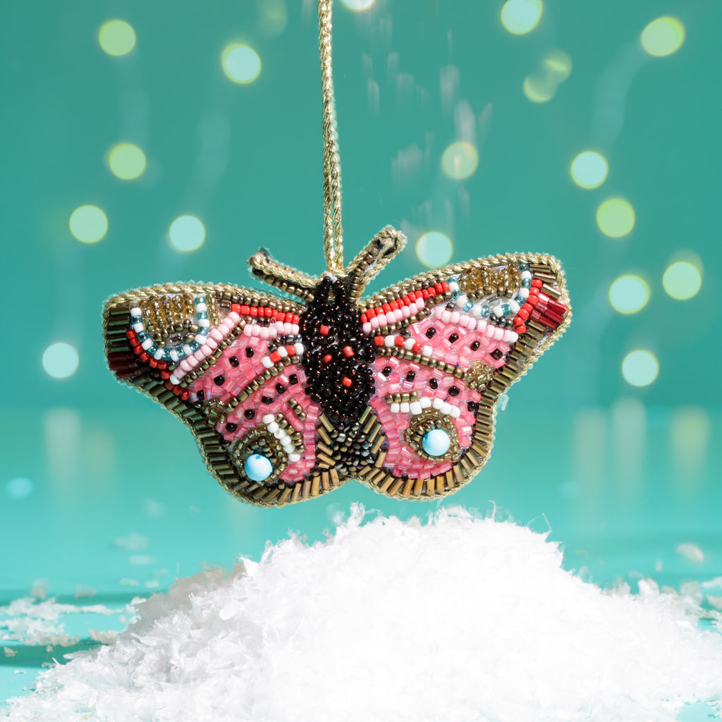 Decorative butterfly ornament with pink and gold details hanging over snow against a teal background