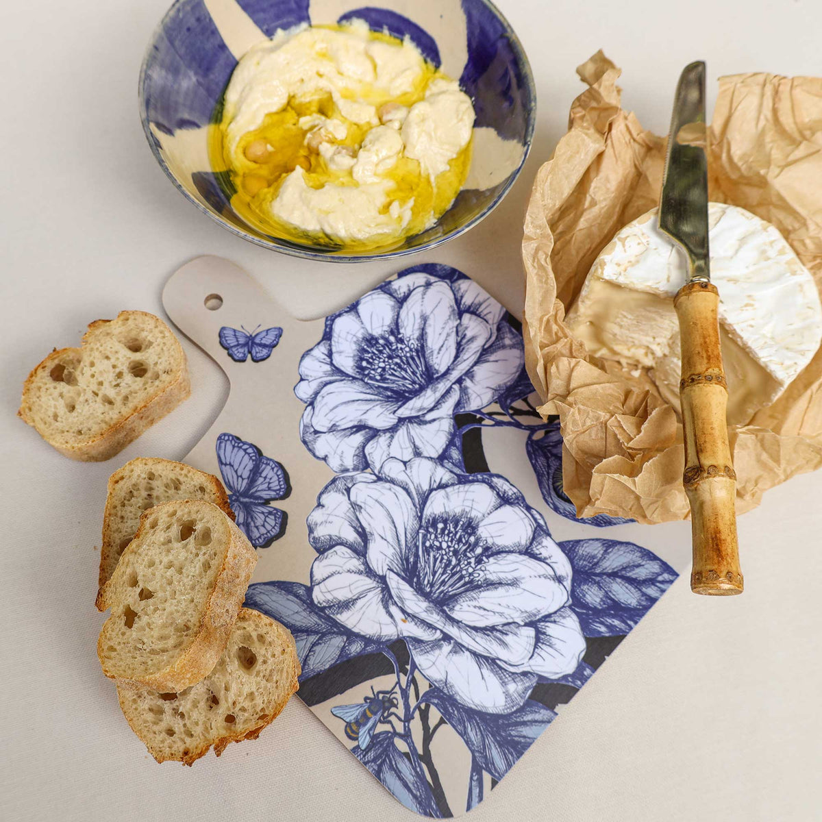 Cream Cornwall floral chopping board featuring elegant blue botanical illustrations, butterflies and a bee, styled with creamy Brie cheese, slices of crusty bread and a bowl of whipped dip drizzled with olive oil on a neutral linen background.