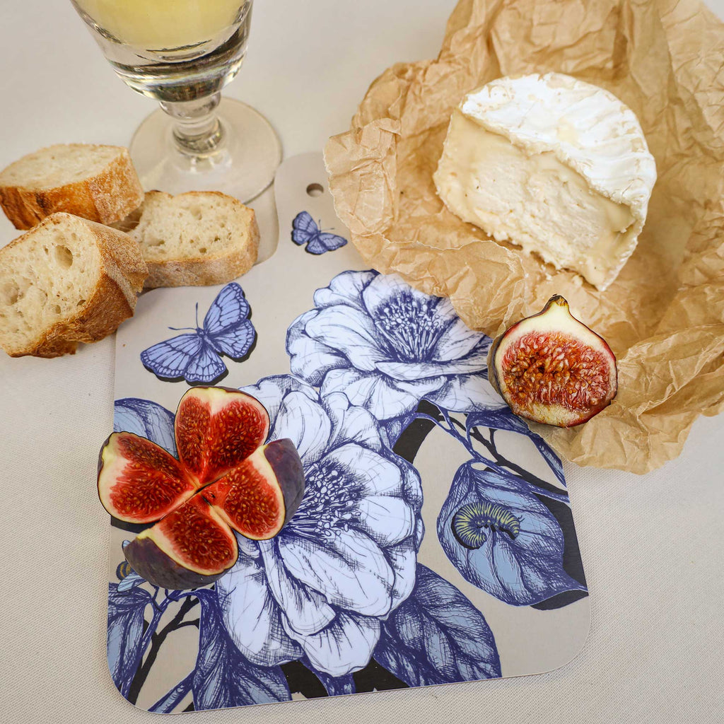 Cream Cornwall floral chopping board featuring intricate blue botanical illustrations and butterflies, styled with fresh figs, creamy Brie cheese, crusty bread and a glass of white wine on a neutral linen tablecloth.