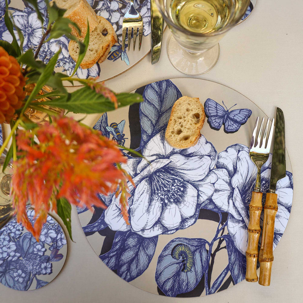 Overhead view of an elegant table setting featuring a round placemat with intricate blue botanical illustrations of large blooming flowers, leaves, a butterfly, a bee and a green caterpillar on a soft beige background. A slice of rustic baguette rests on the placemat beside bamboo-handled cutlery, with a glass of white wine above. Vibrant orange and coral flowers, lush greenery and a matching floral coaster complete the arrangement on a cream linen tablecloth.