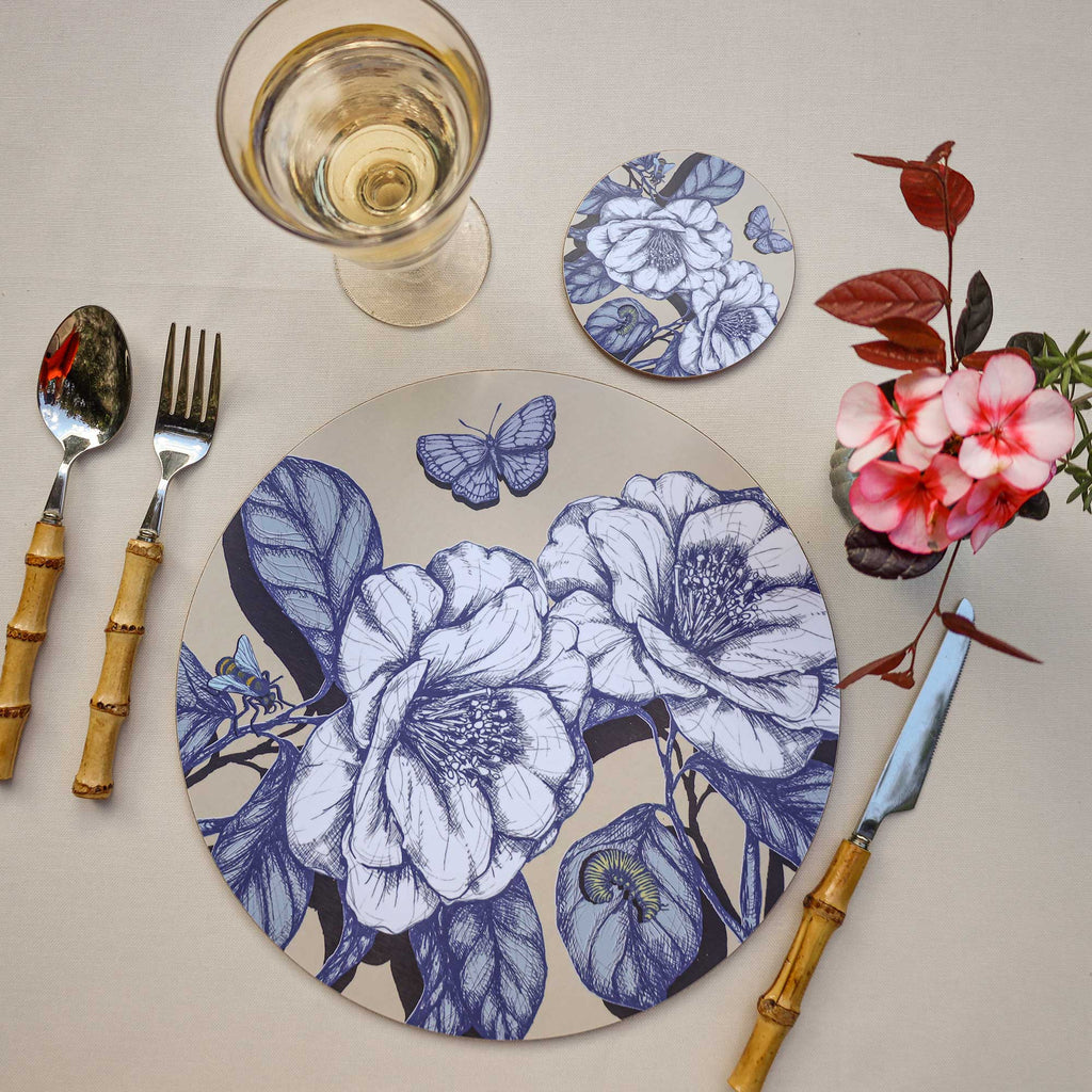 Cream Cornwall round botanical placemat and matching coaster featuring elegant blue and white floral illustrations, butterflies and a bee on a soft cream background, styled with bamboo-handled cutlery, a glass of white wine and delicate pink flowers on a neutral tablecloth.