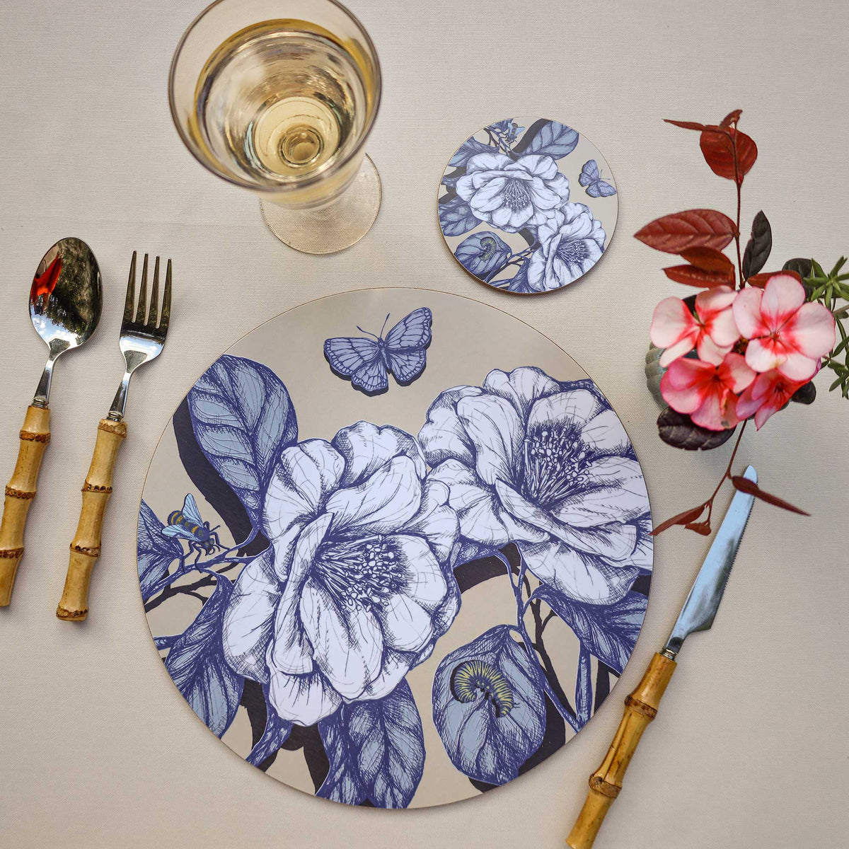 Overhead view of an elegant table setting featuring a round placemat with intricate blue botanical illustrations of large blooming flowers, leaves, a butterfly, a bee and a caterpillar on a soft beige background. A matching floral coaster, glass of white wine and bamboo-handled cutlery complement the design, while delicate pink flowers and burgundy foliage add a vibrant touch against the cream linen tablecloth.