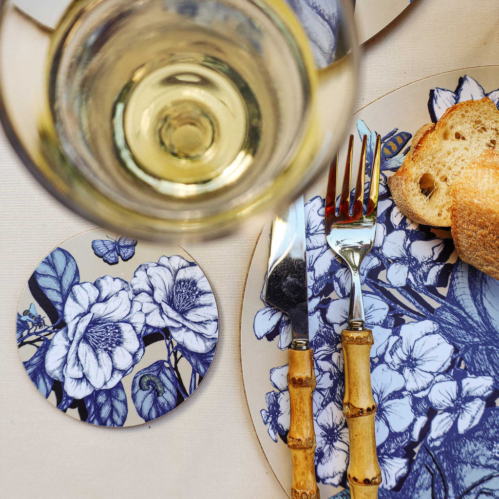 Cream Cornwall botanical coaster featuring elegant blue and white floral illustrations and delicate butterflies, styled beside a matching floral placemat, bamboo-handled cutlery, crusty bread and a glass of white wine on a cream tablecloth.