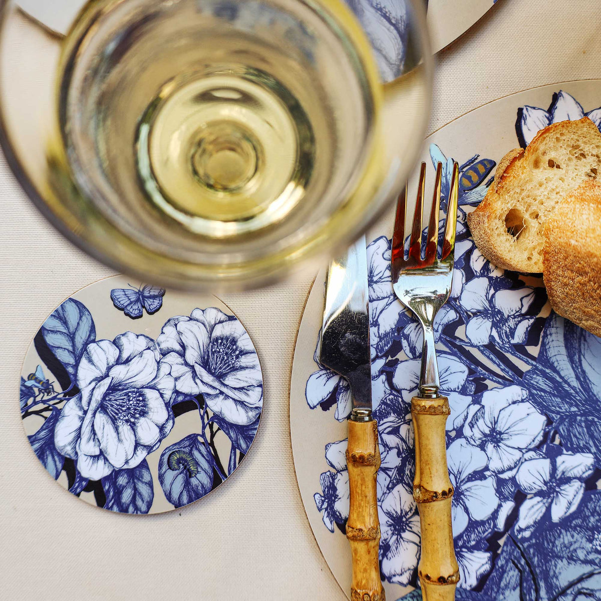Cream Cornwall botanical coaster featuring elegant blue and white floral illustrations and delicate butterflies, styled beside a matching floral placemat, bamboo-handled cutlery, crusty bread and a glass of white wine on a cream tablecloth.