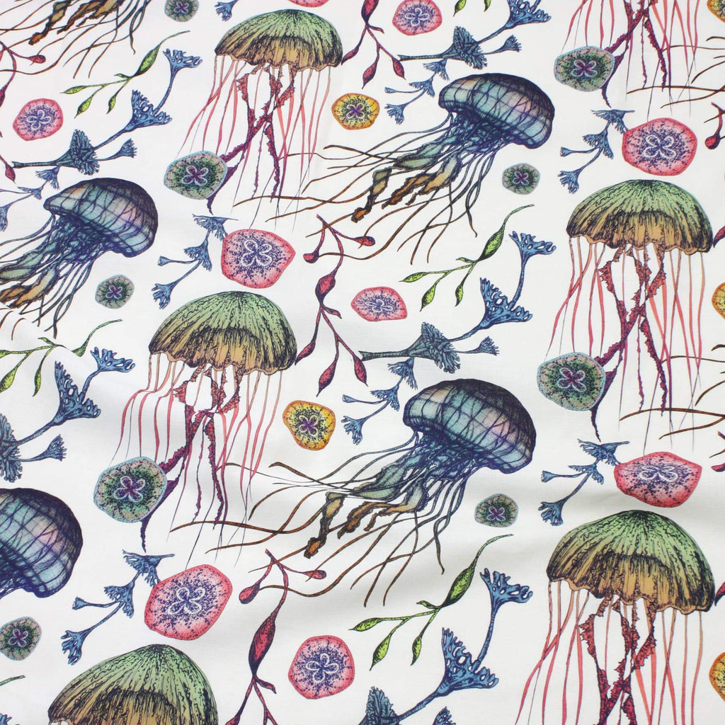 A vibrant flat lay of Cream Cornwall’s Canyons Reef fabric, showcasing a marine-inspired pattern on an off-white background. The design features hand-drawn jellyfish with green and blue domes and flowing tentacles in pinks and purples, surrounded by delicate seaweed, coral-like flora, and sea anemone motifs in soft greens, blues, and pinks. The arrangement captures the movement and wonder of an underwater world.