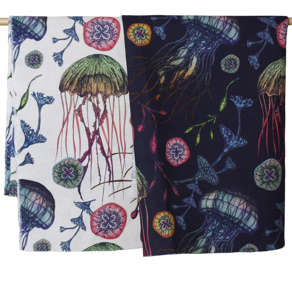 Two hanging fabric panels from Cream Cornwall’s Canyons Reef collection, showcasing a vivid underwater design of jellyfish, seaweed, and marine flora. The left fabric features a crisp off-white background with colourful illustrations in shades of green, red, and blue. The right fabric has the same pattern on a deep navy background, giving the marine motifs a more dramatic, nocturnal appearance. Both fabrics are draped over a wooden rod for display.
