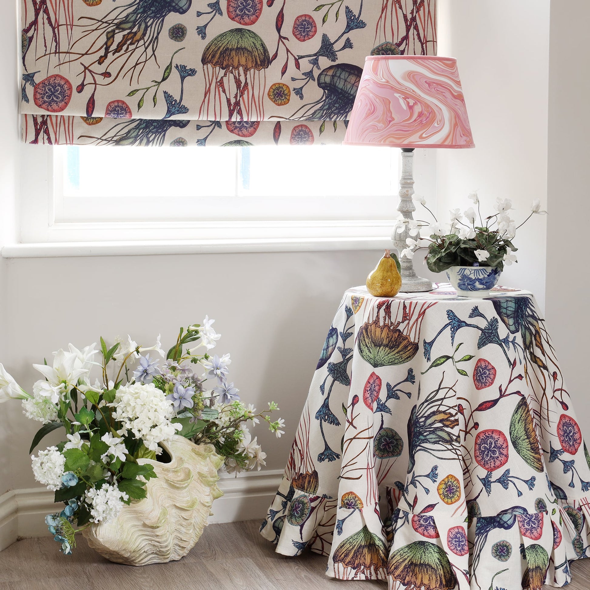 A coastal-inspired interior featuring Cream Cornwall’s Canyons Reef fabric in an off-white and natural colourway, used for both a Roman blind and a round tablecloth with a ruffled edge. The fabric showcases a detailed marine pattern of jellyfish, coral, and sea plants in soft greens, blues, reds, and pinks. A pink marbled lampshade, decorative ceramic pear, and fresh white flowers in a clam shell planter complete the serene, ocean-themed setting.