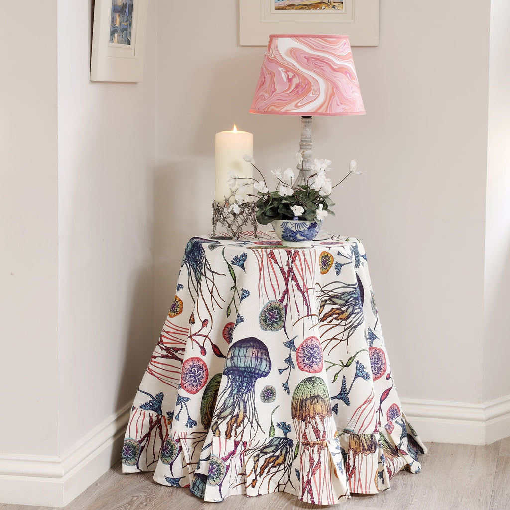 A charming corner setup featuring a round table dressed in Cream Cornwall’s Canyons Reef fabric in off-white, adorned with vivid illustrations of jellyfish, seaweed, and underwater flora. The table is topped with a marbled pink lampshade, a blue and white floral pot holding white cyclamen flowers, and a lit pillar candle in a coral-inspired holder. The light and coastal-themed space is finished with soft wall colours and framed artwork, evoking a tranquil seaside ambiance.