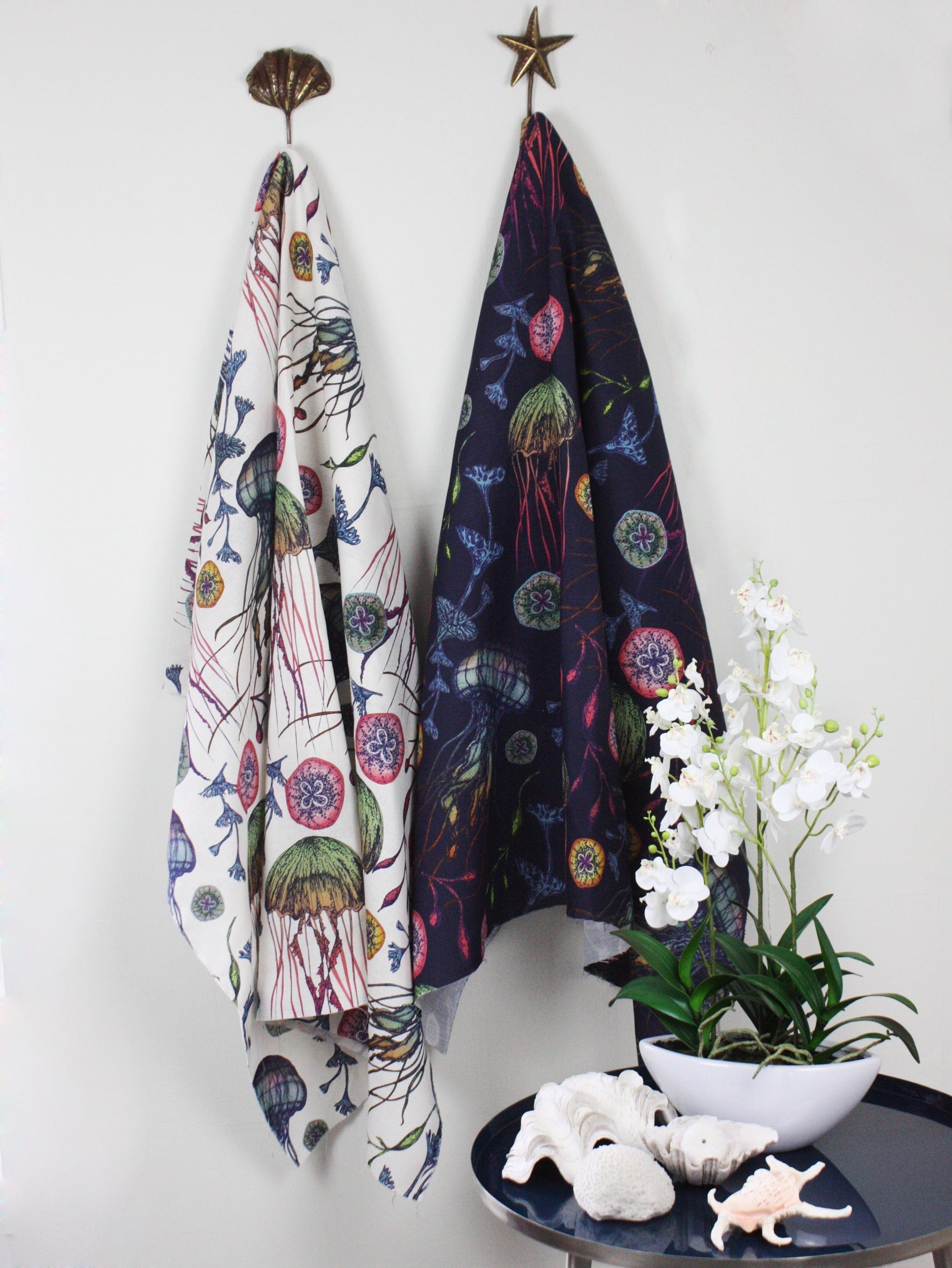 Two draped fabrics from Cream Cornwall’s Canyons Reef collection hang side by side on decorative brass hooks—a shell and a star. The left fabric features an off-white background with colourful illustrations of jellyfish, seaweed, and sea creatures, while the right fabric has the same vibrant design set against a deep navy background. Beneath them is a small round table with a white orchid plant in a bowl, coral decor, and sea-inspired shells, enhancing the coastal aesthetic of the display.