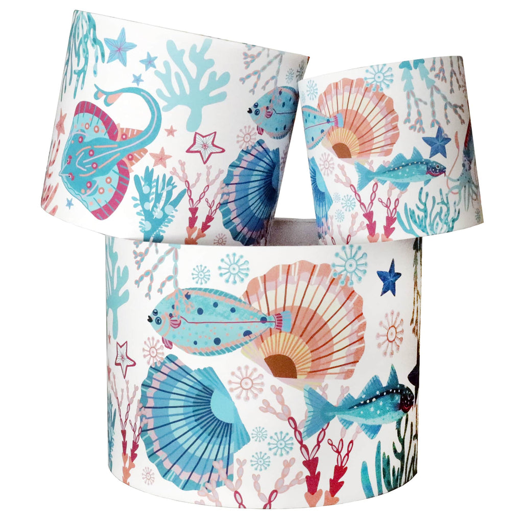 A set of three cylindrical lampshades featuring a vibrant coastal design. The pattern includes colorful sea creatures like fish and stingrays, alongside coral, starfish, and seashells in shades of blue, pink, and orange on a white background. Each lampshade showcases a unique section of the underwater-themed artwork, adding a lively and nautical touch to home décor.