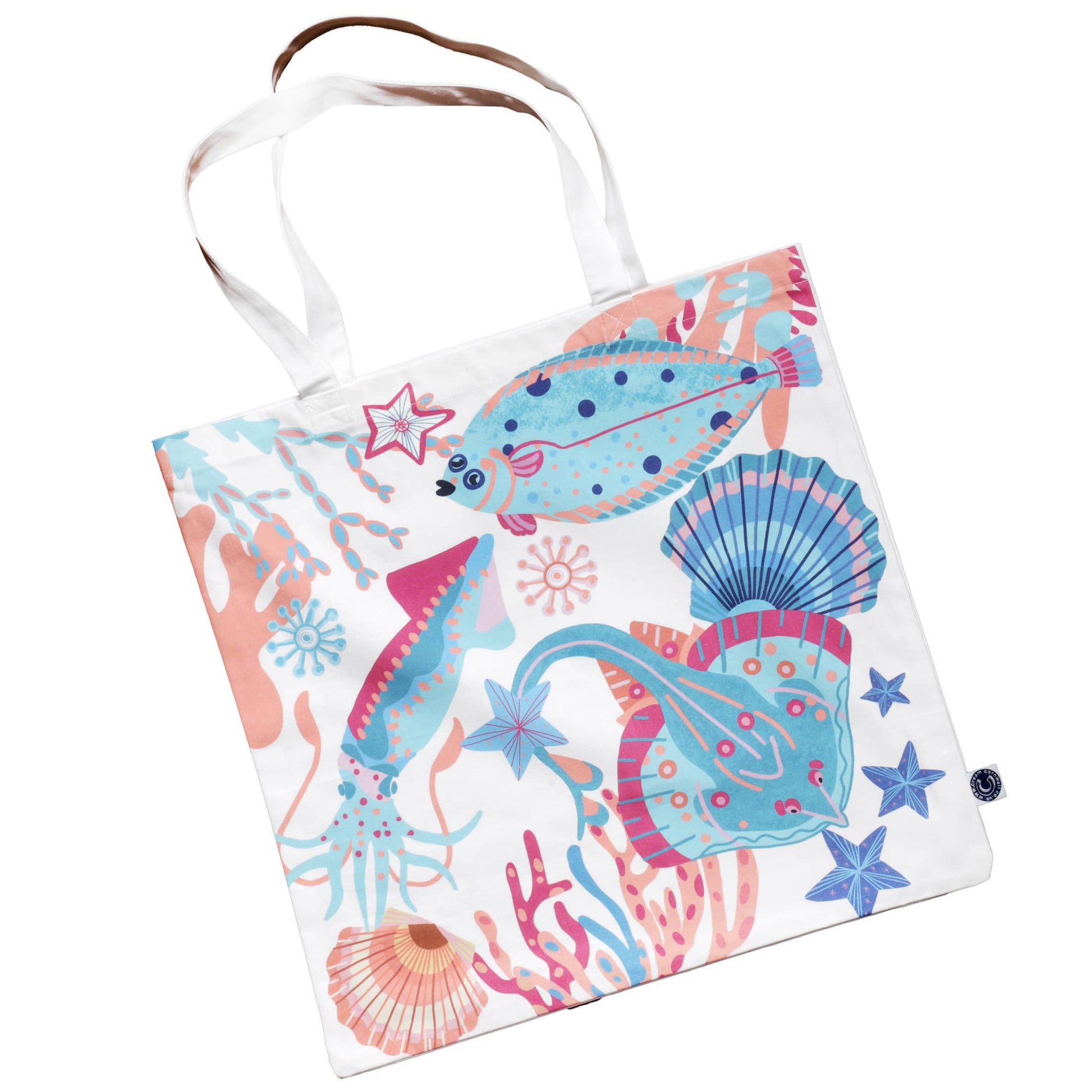 Fabric tote bag featuring a colorful marine-themed print with illustrations of a flat fish, squid, skate, seashells, coral, and starfish. The design is in shades of turquoise, pink, and blue on a white background, giving it a fun and vibrant ocean-inspired look. The bag has sturdy white handles.