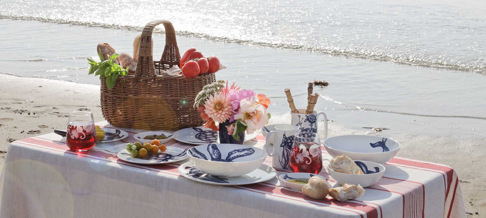 A styled beach picnic setup on a white and red striped tablecloth with blue-and-white ceramic dishes, a wicker picnic basket, flowers, bread, and red tomatoes, set against a sandy shoreline and ocean water.