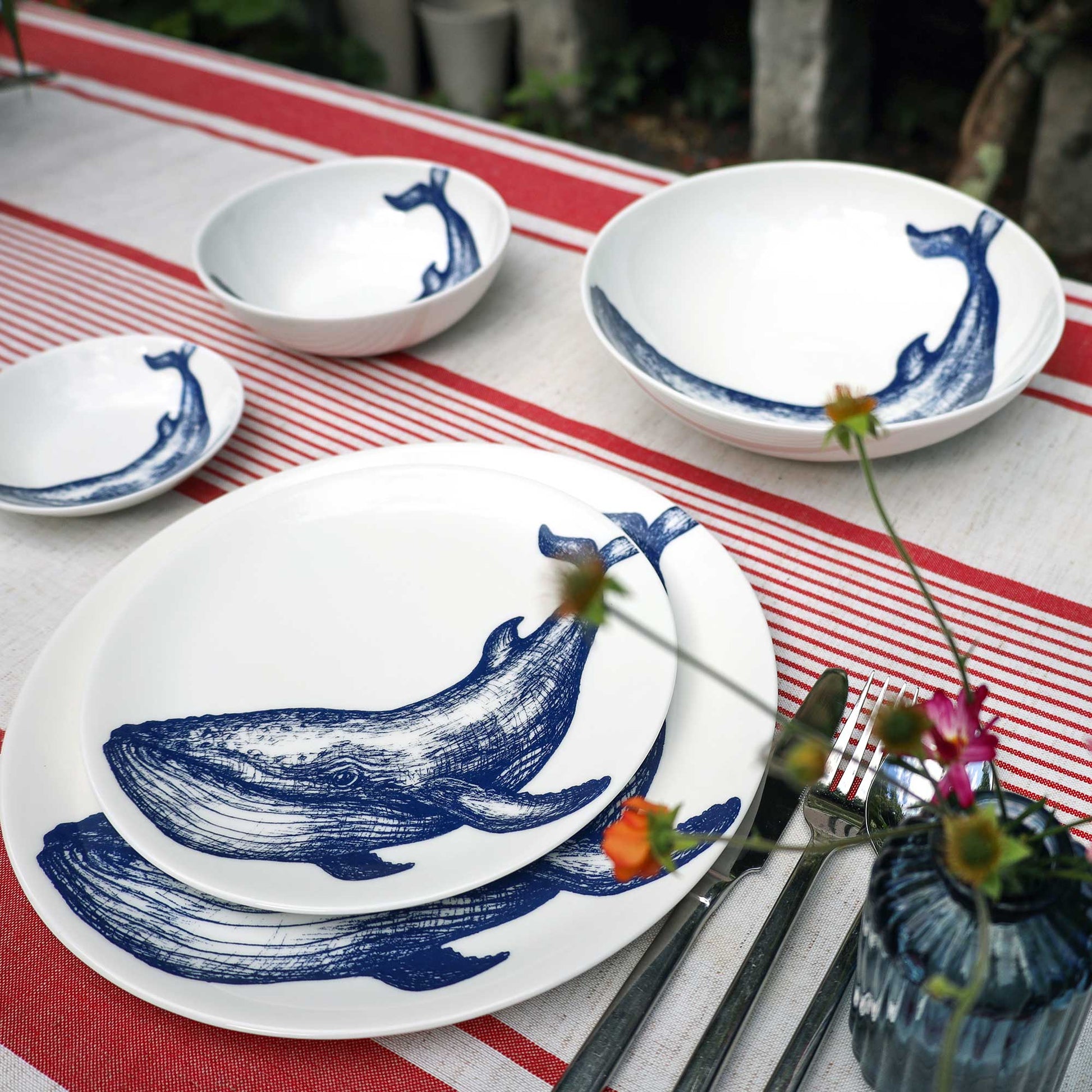 A set of white ceramic tableware featuring blue whale illustrations, arranged on a red and white striped table runner with silver cutlery and a small blue glass vase with flowers.