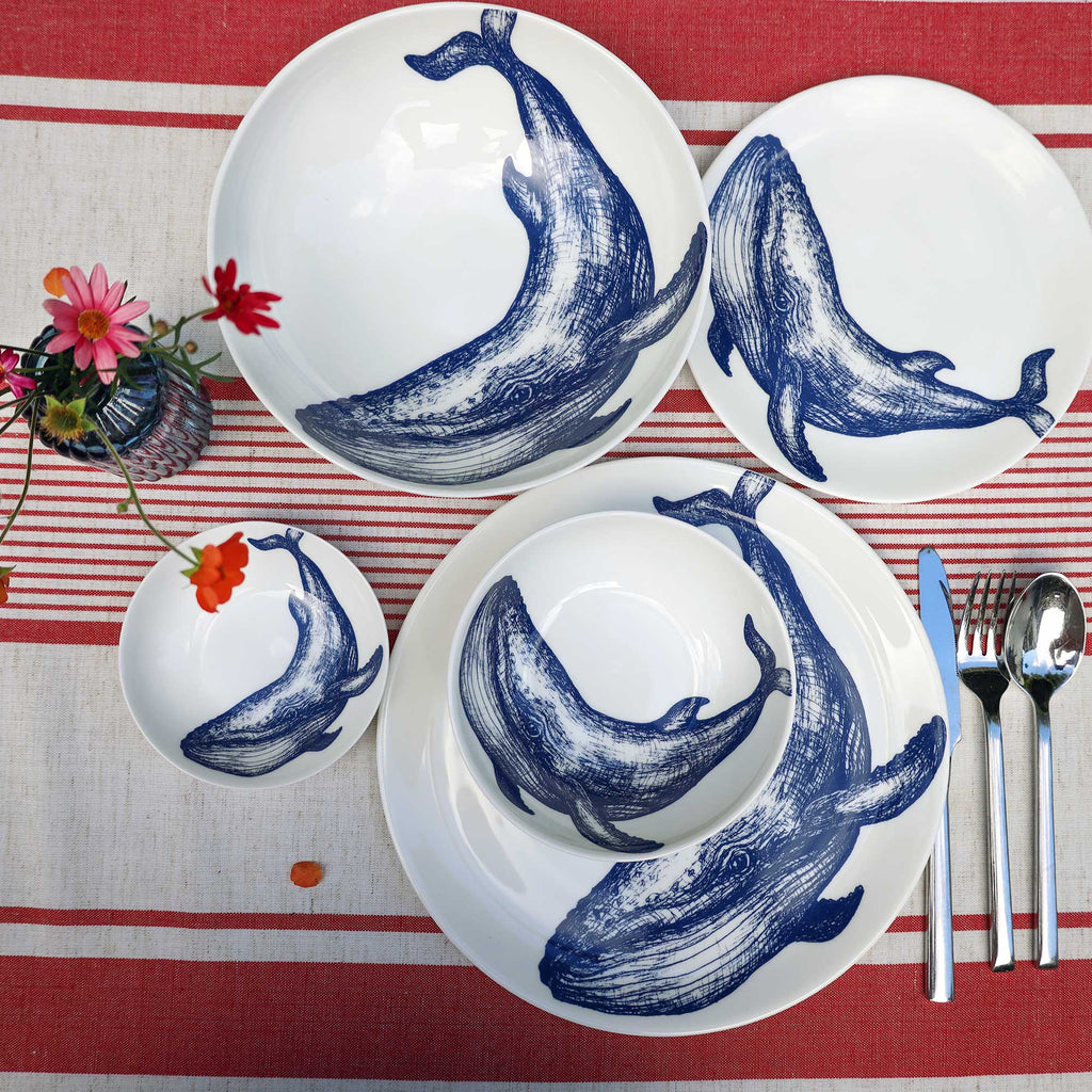 A set of white ceramic dinnerware featuring blue whale illustrations, arranged on a red and white striped tablecloth with silver cutlery and a small floral arrangement.