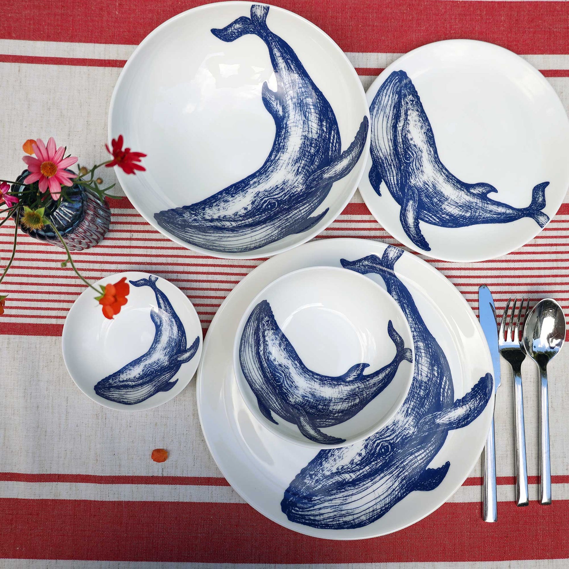 A set of white ceramic dinnerware featuring blue whale illustrations, arranged on a red and white striped tablecloth with silver cutlery and a small floral arrangement.